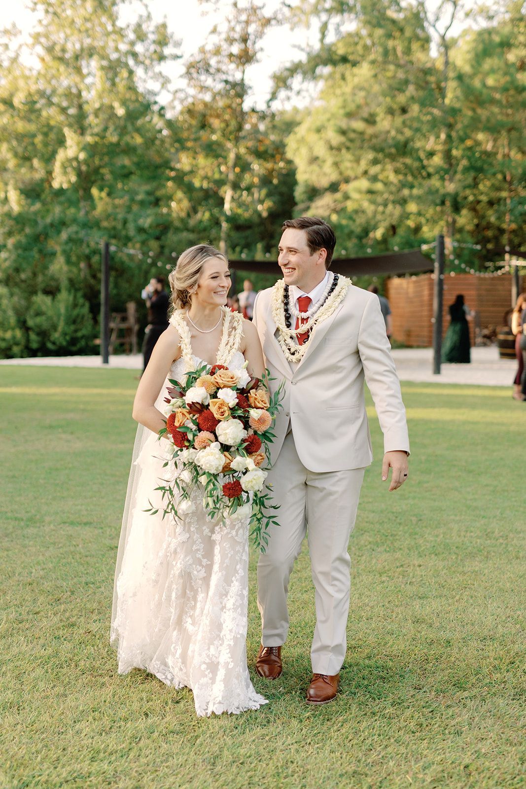 Bride and groom walking hand in hand on grass, smiling in a garden setting.