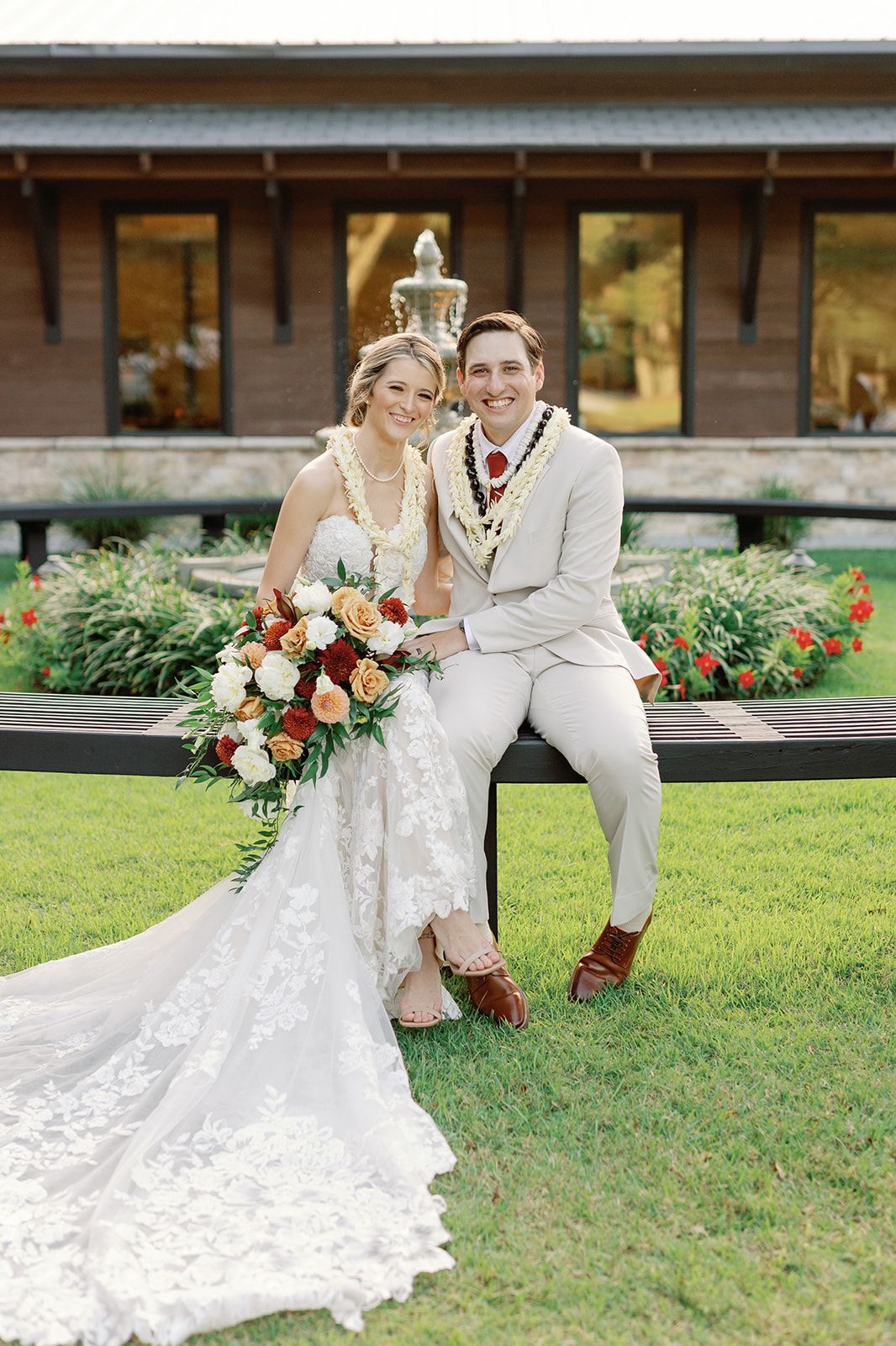 Two wedding guests posing outdoors, one in a white dress and one in a light suit, on a lawn before a building