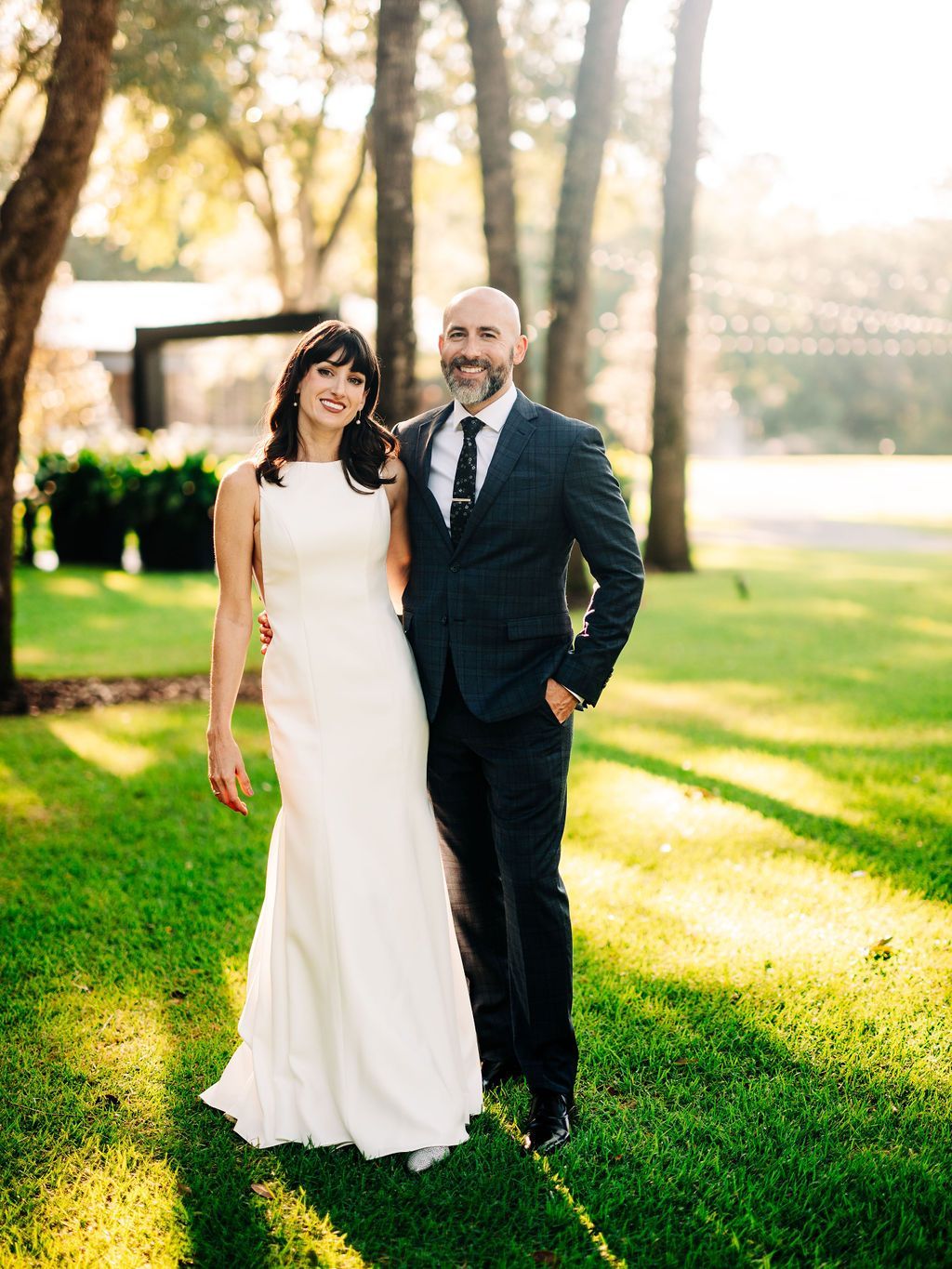 Couple in wedding attire standing on a sunlit lawn with trees in the background