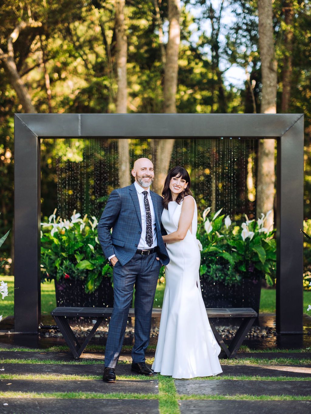 Couple posing outdoors in wedding attire under a large metal frame, with trees and flowers behind them.