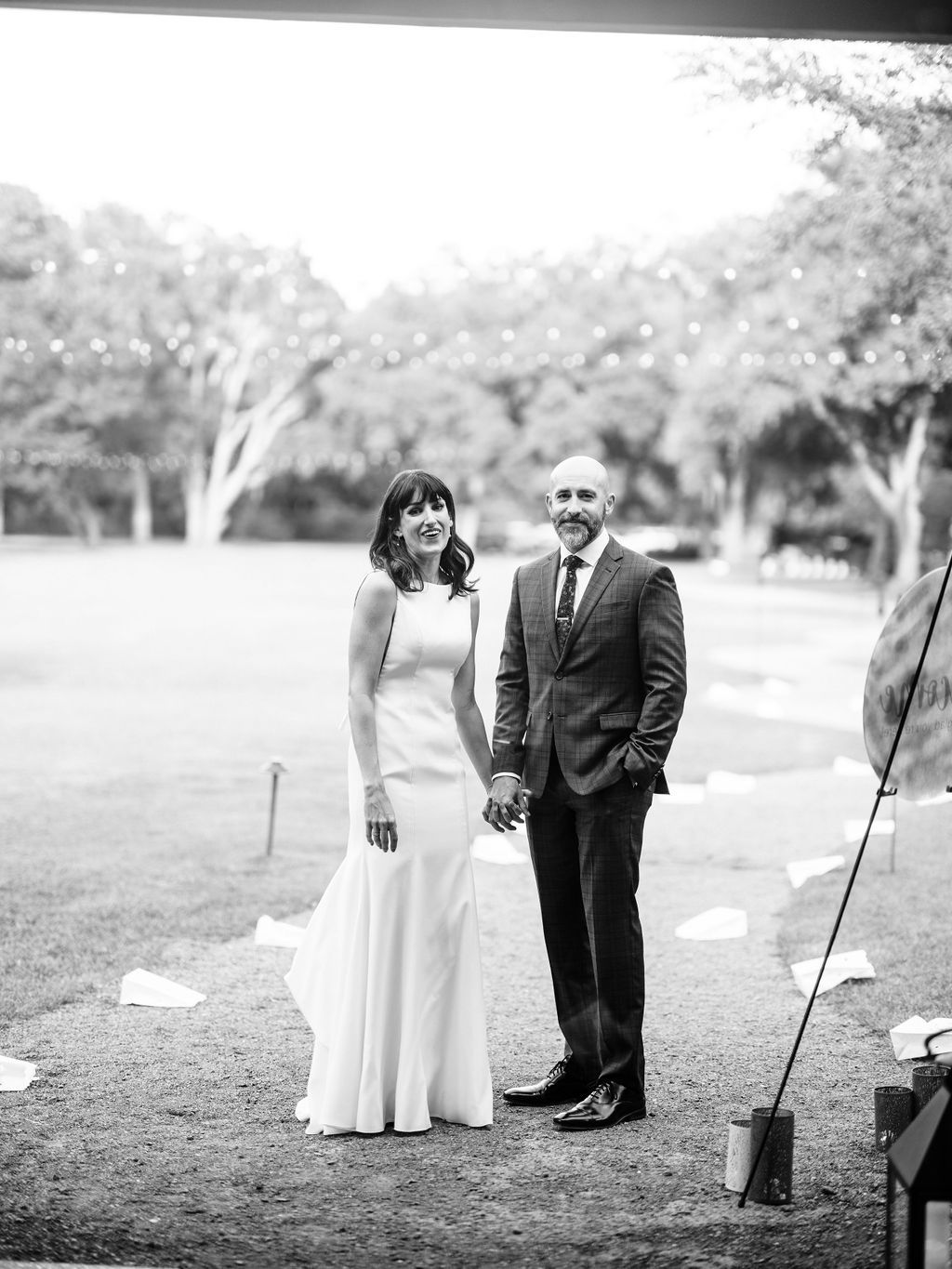 Bride and groom standing outdoors on a lawn, holding hands, in a black-and-white wedding photo
