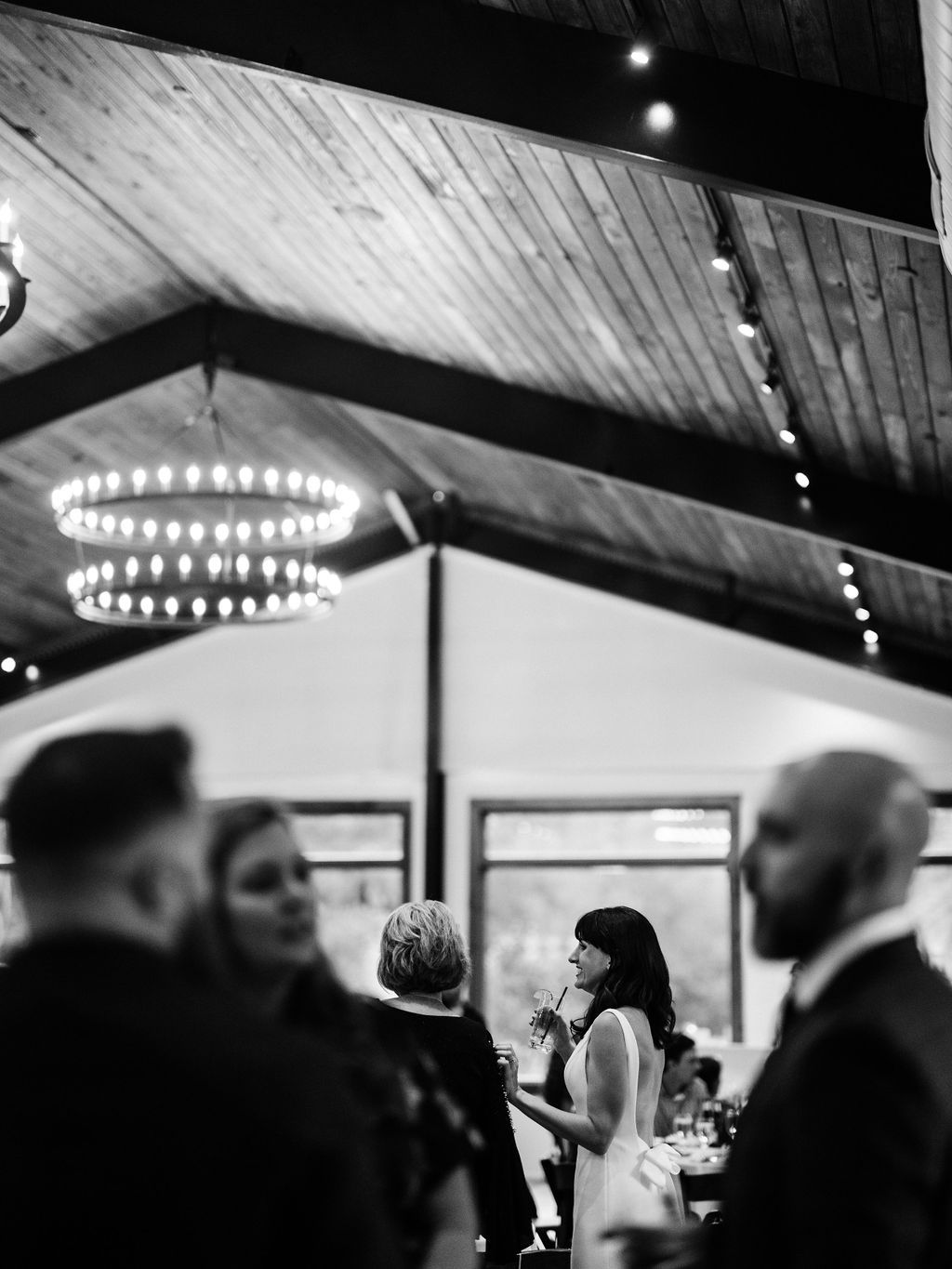 Black-and-white indoor event scene with blurred guests under a high wooden ceiling and chandelier lights