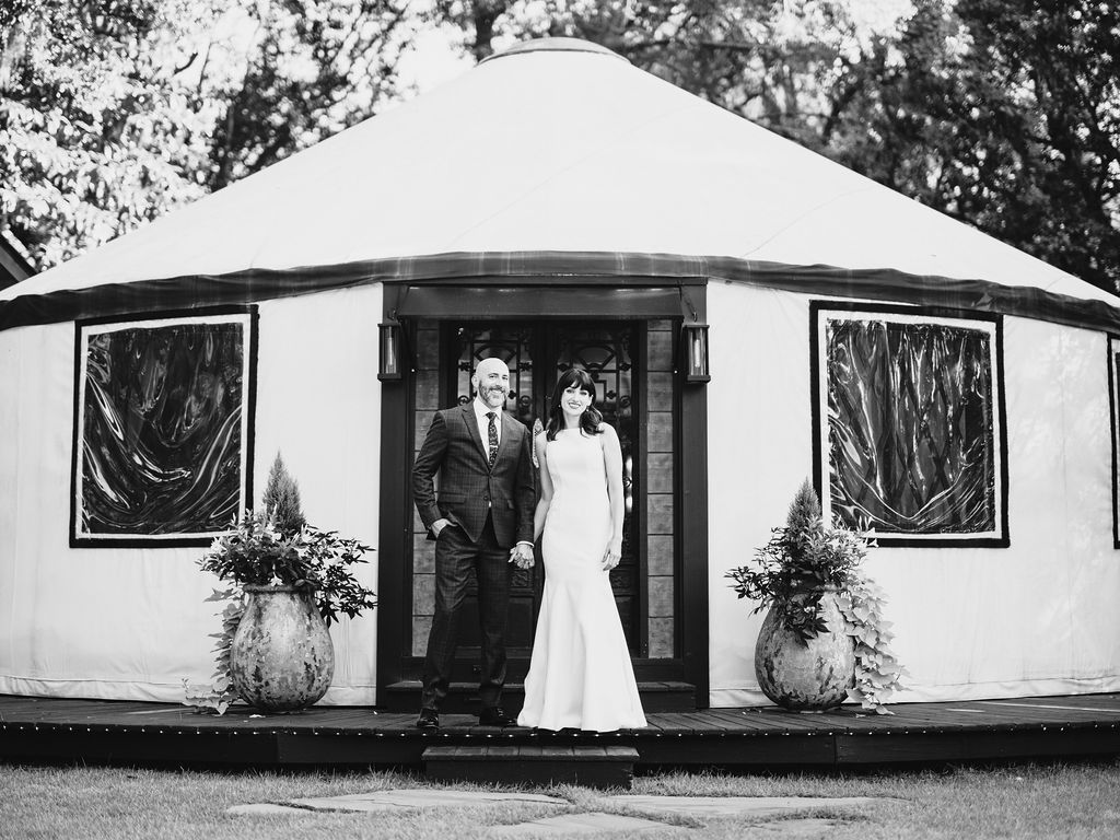 Bride and groom stand at the entrance of a white tent, framed by potted plants.