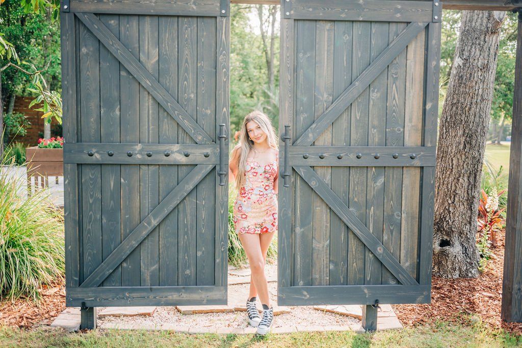 Woman in a floral dress standing between open wooden gates in a garden.