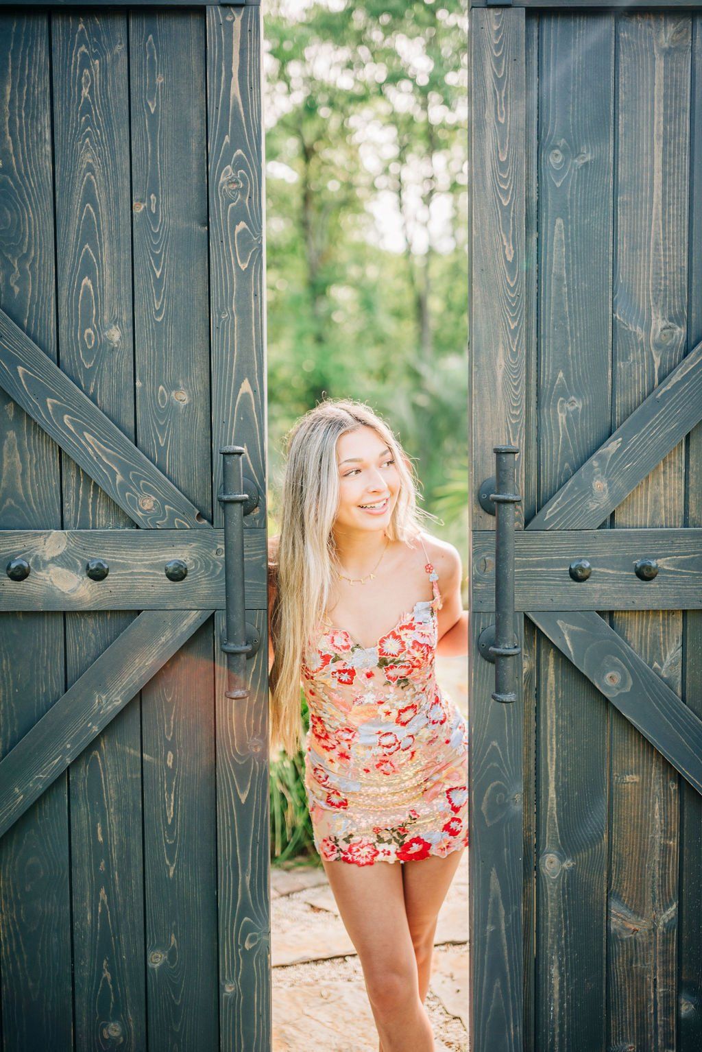 Woman in a floral dress standing between open wooden gates on a sunlit path.
