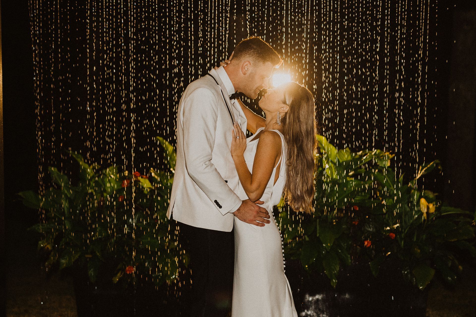 Couple kissing in white attire beneath string lights at a nighttime celebration