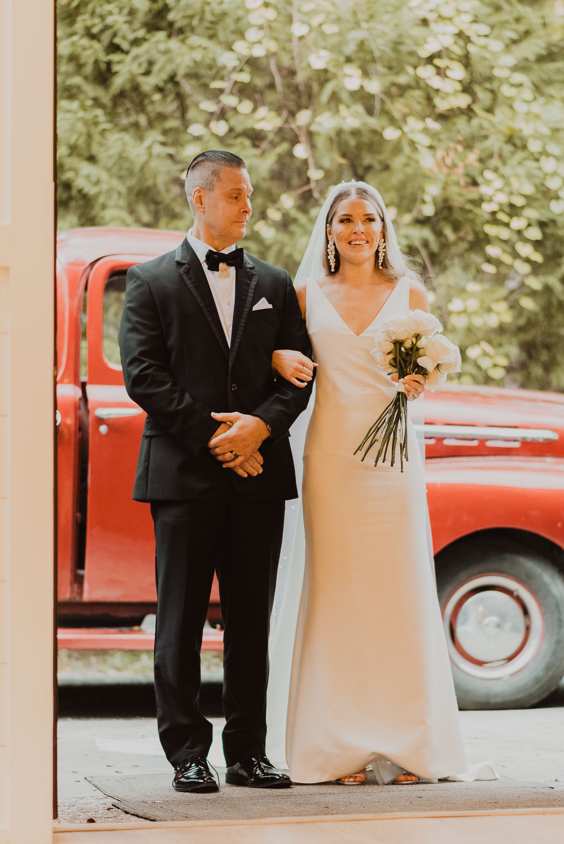 Bride and groom standing together outdoors beside a red vintage truck