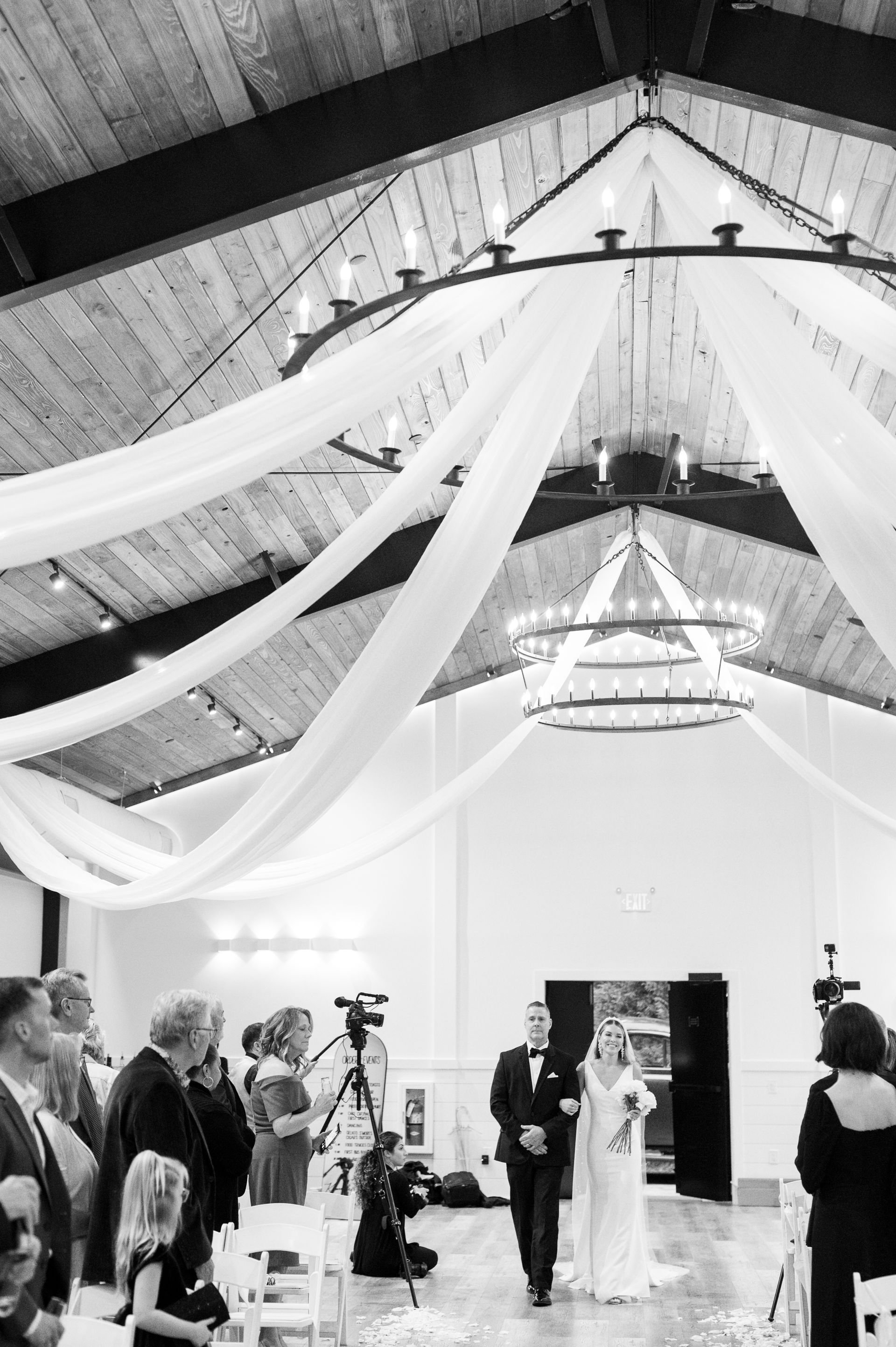 Bride standing under draped ceiling fabric in a decorated hall, with guests and photographers watching.
