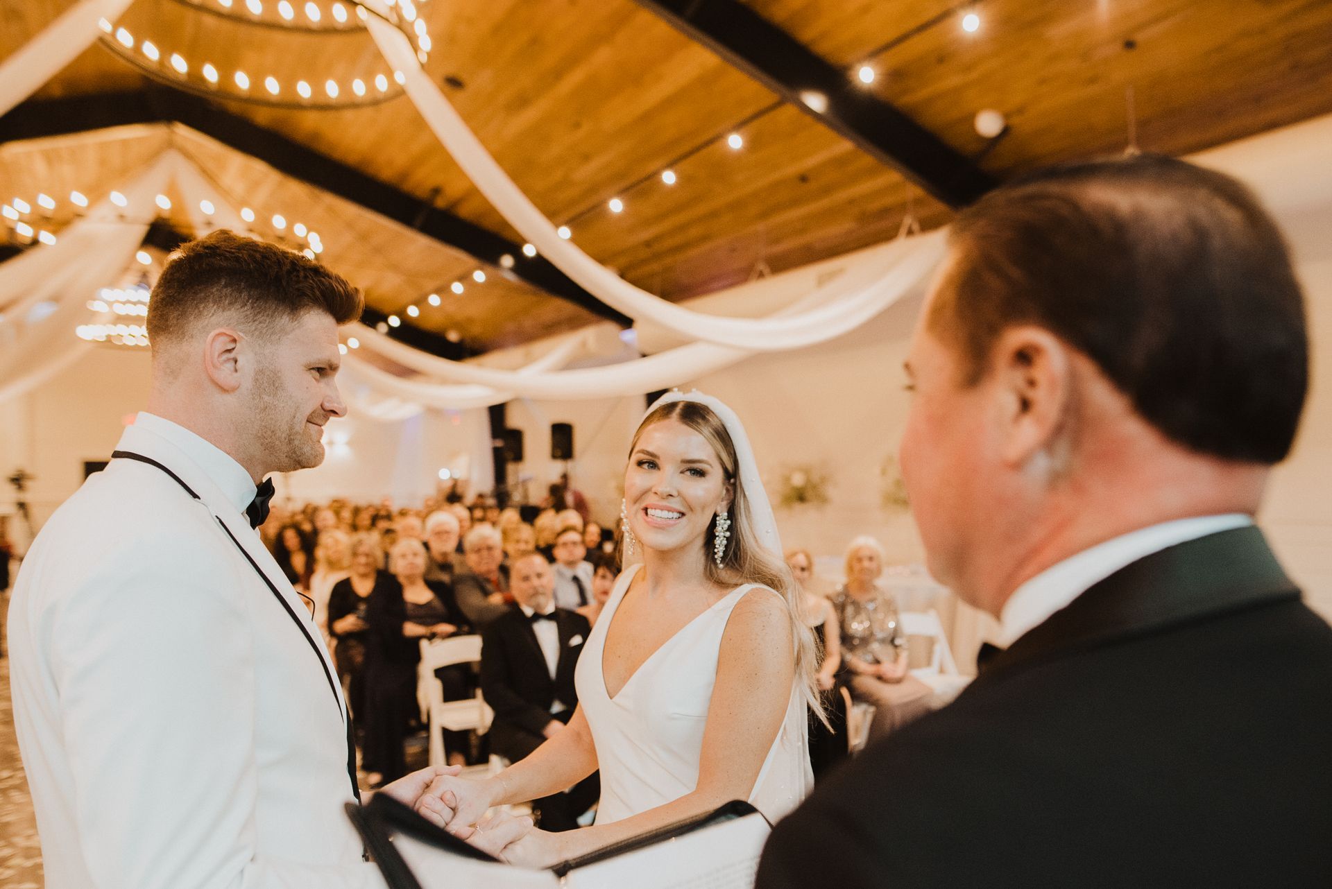 Bride and groom exchanging vows in a decorated wedding hall, smiling as guests watch