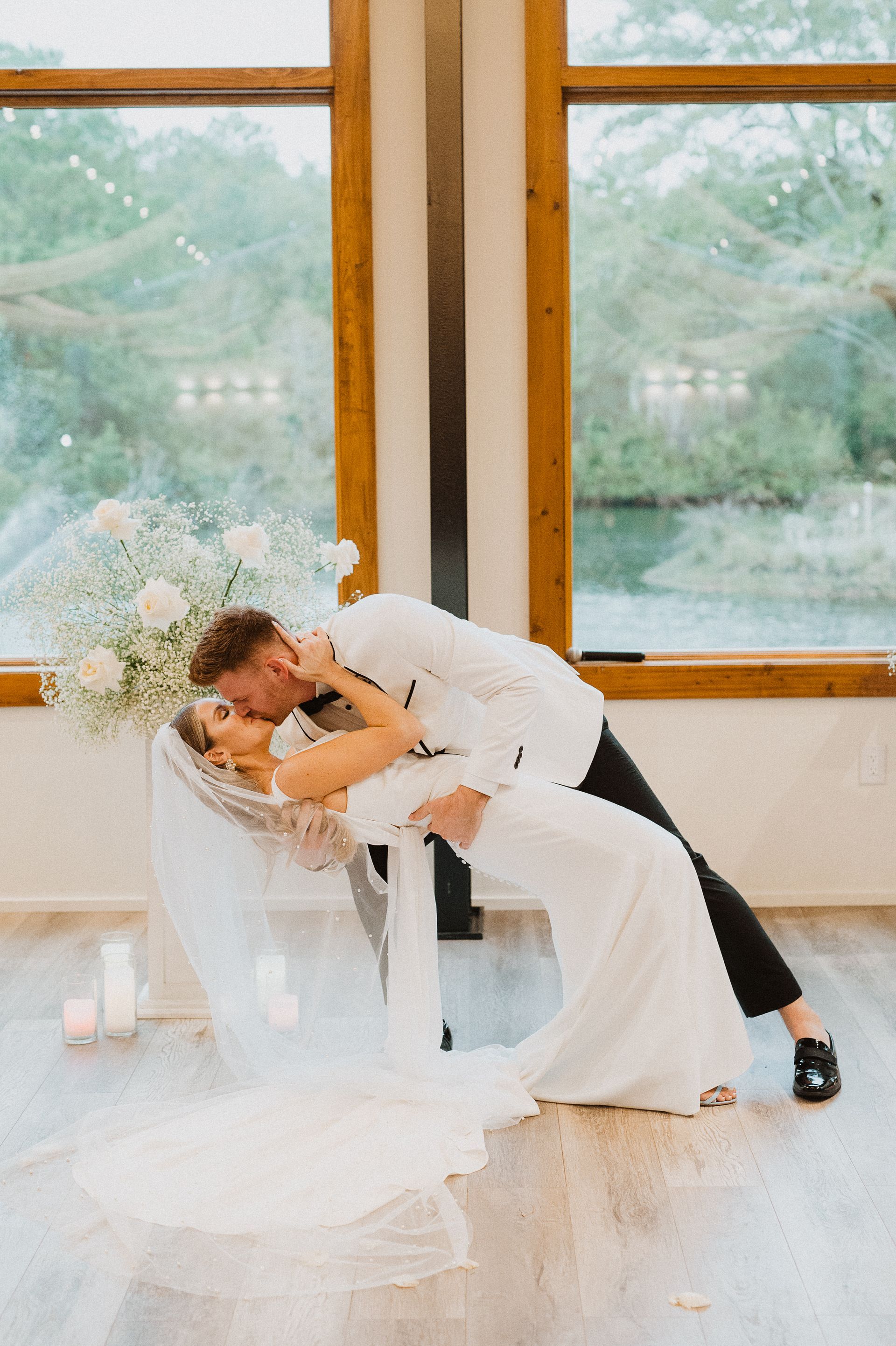 Two people in white outfits embracing in a bright room by large windows, with flowing fabric and flowers nearby