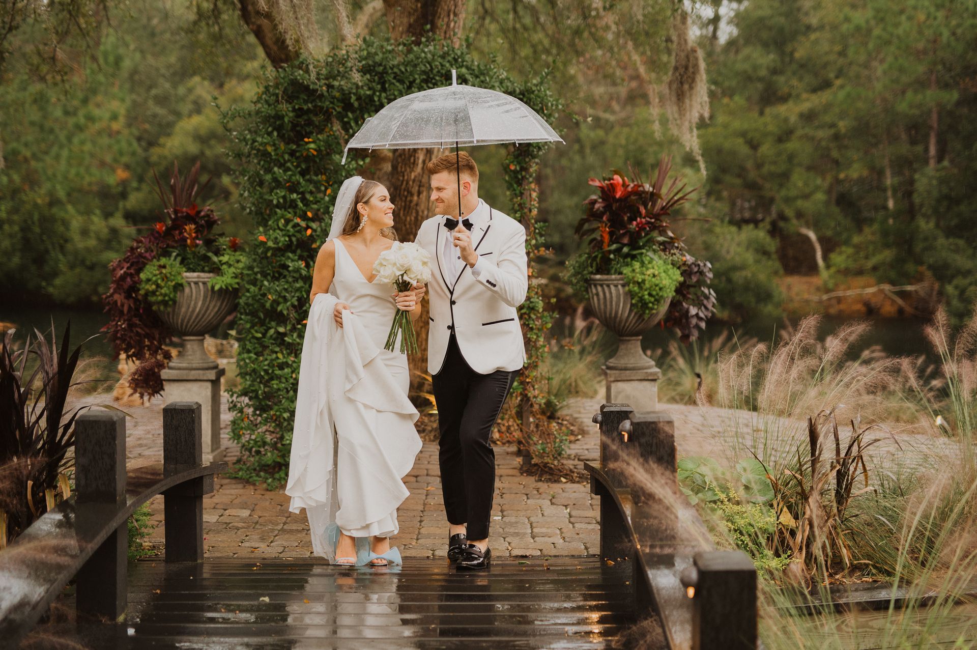 Bride and groom walk under a white umbrella on a wet garden bridge