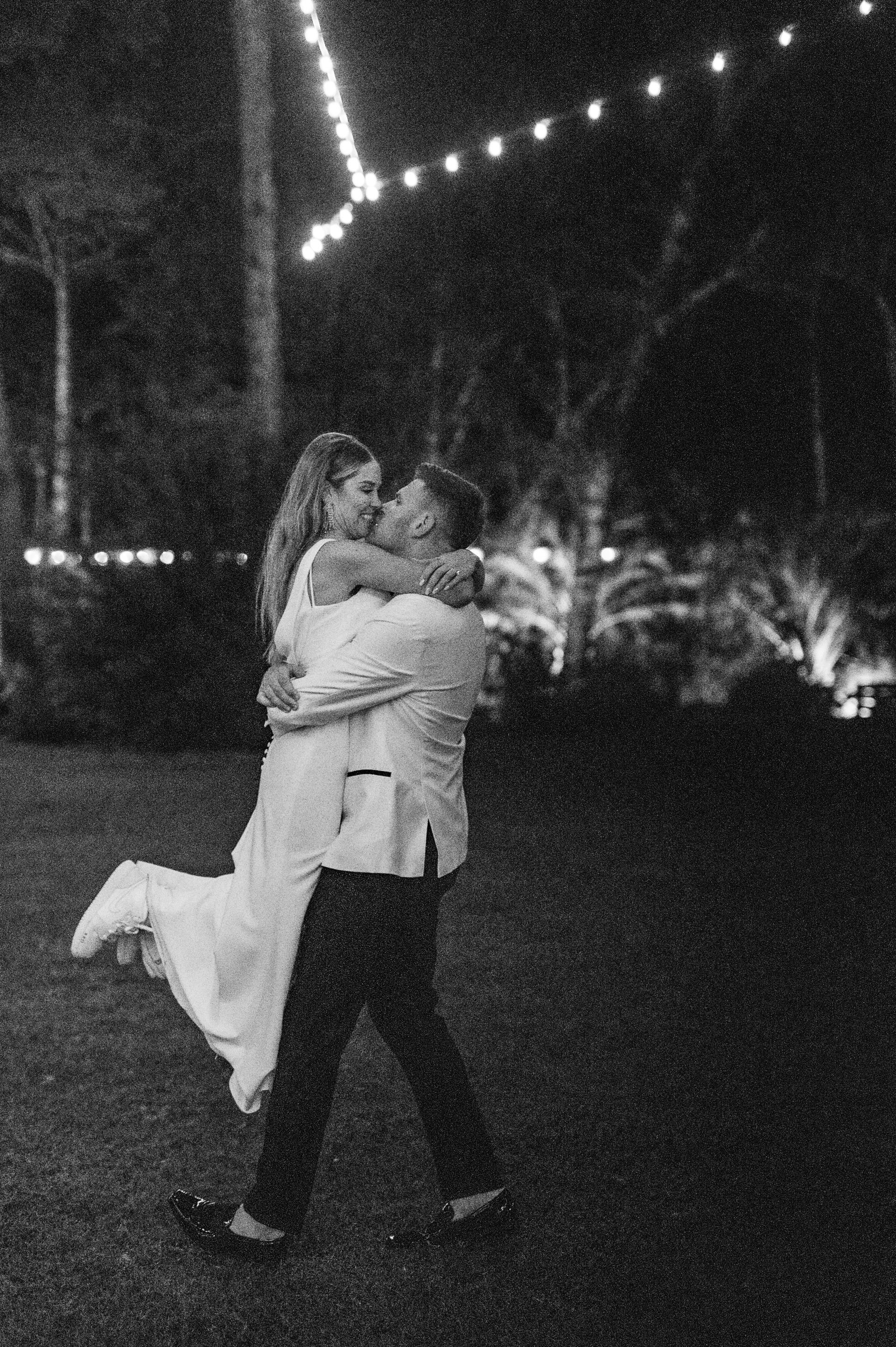 Two people embracing outdoors at night, with string lights and trees in the background.