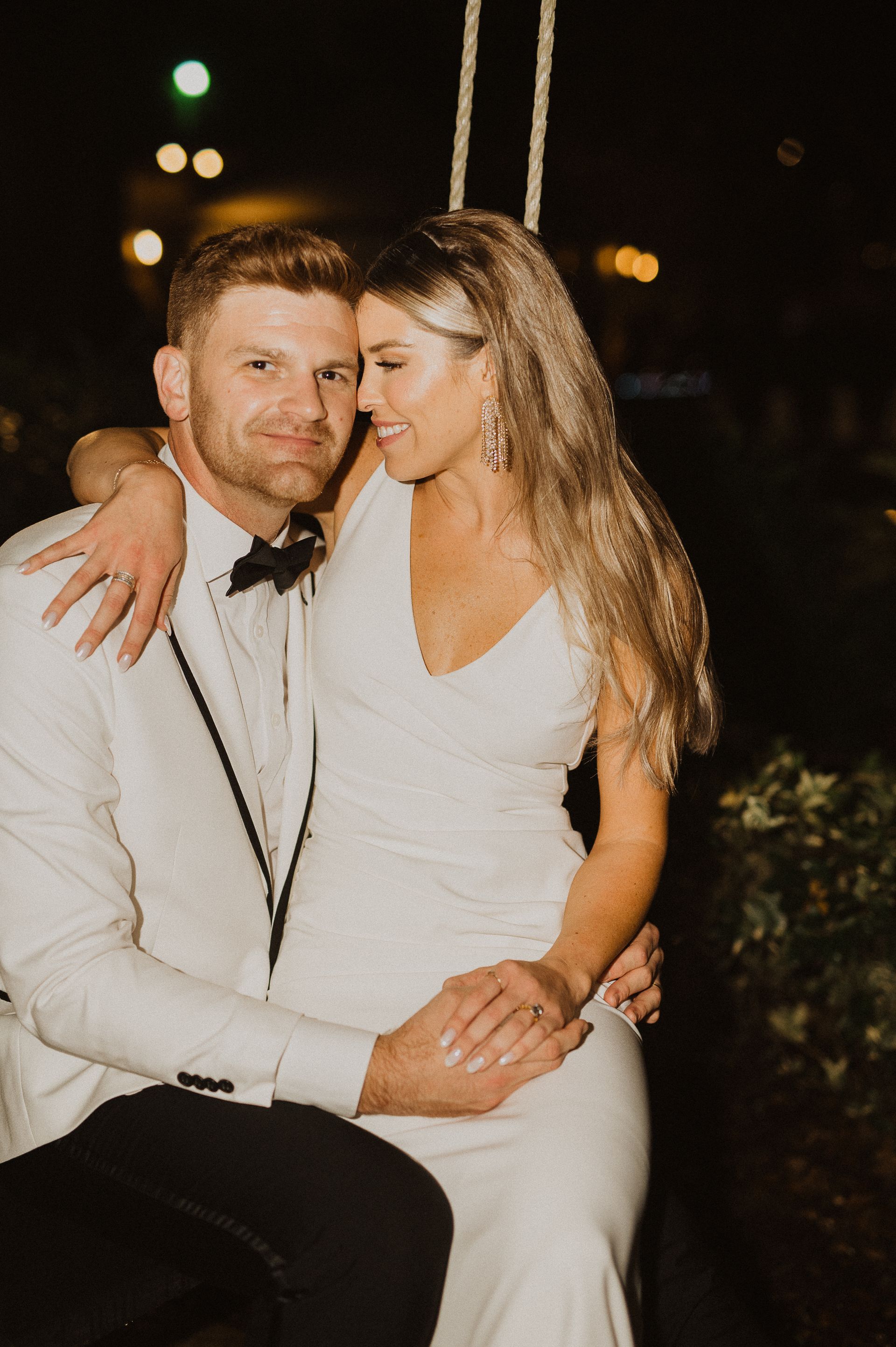 Couple in formal white outfits posing closely outdoors at night, smiling softly.