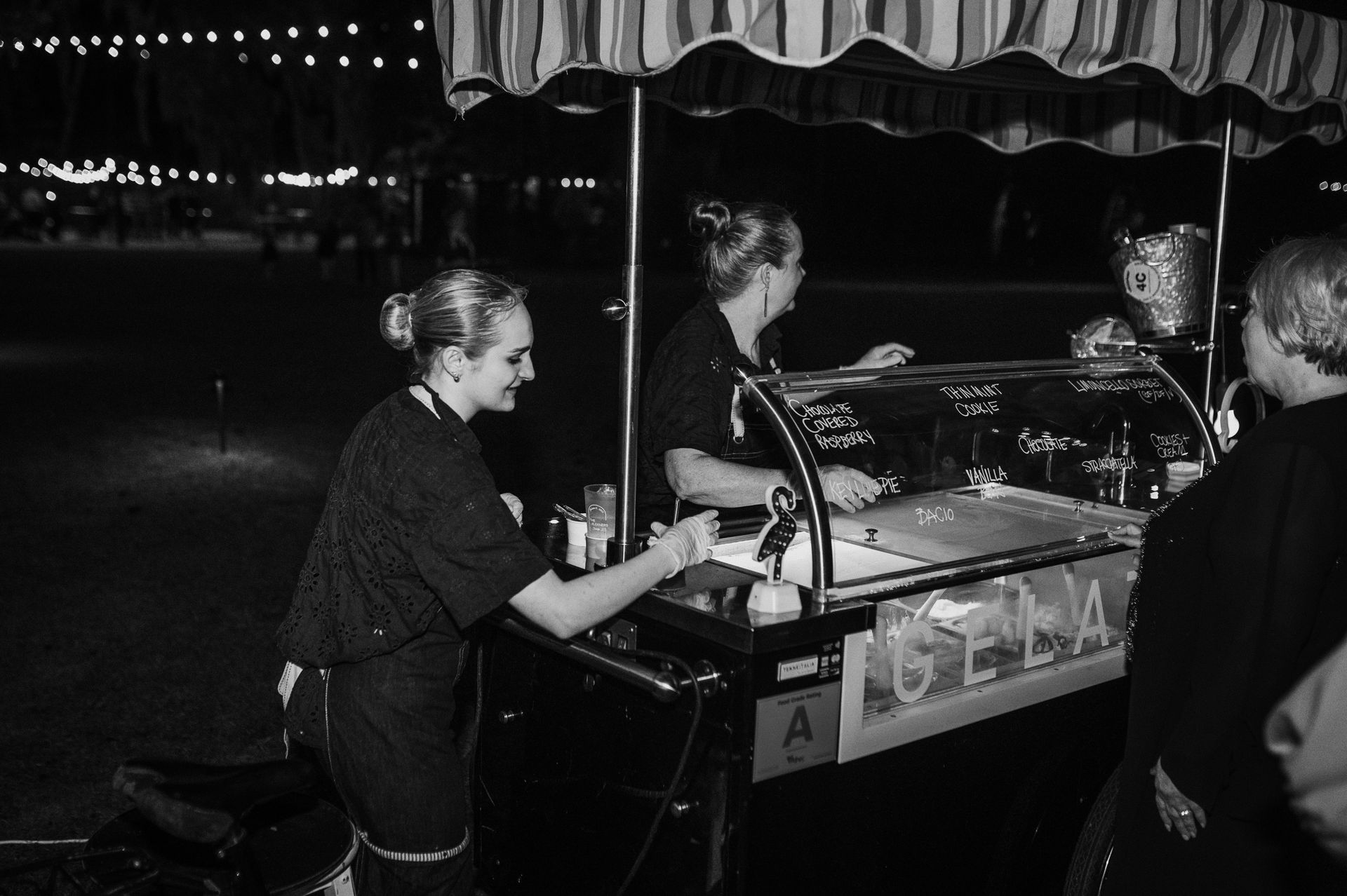 People at a nighttime market stall under striped awnings, serving food and drinks.