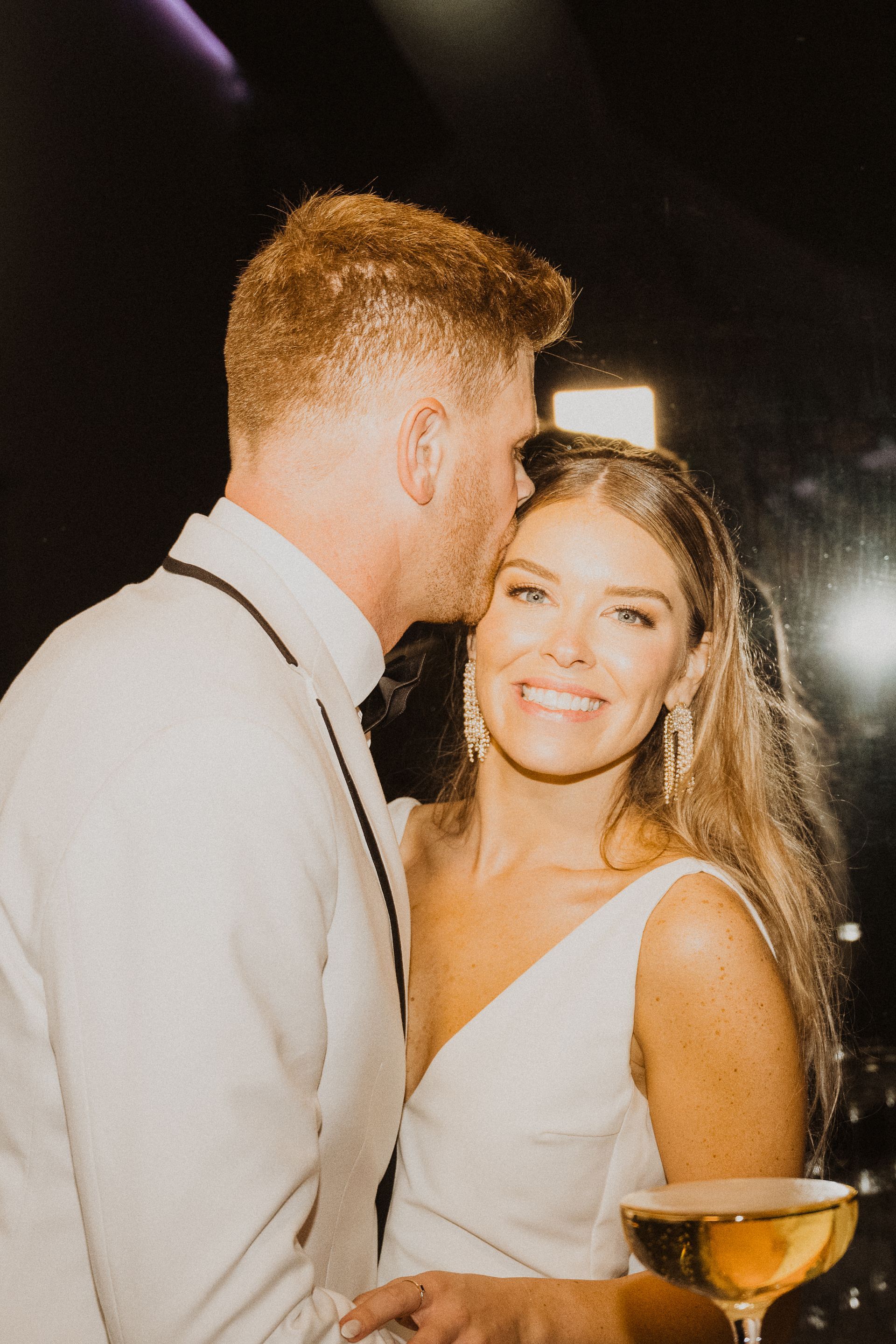 Couple at a dimly lit event, smiling and embracing with a drink in the foreground
