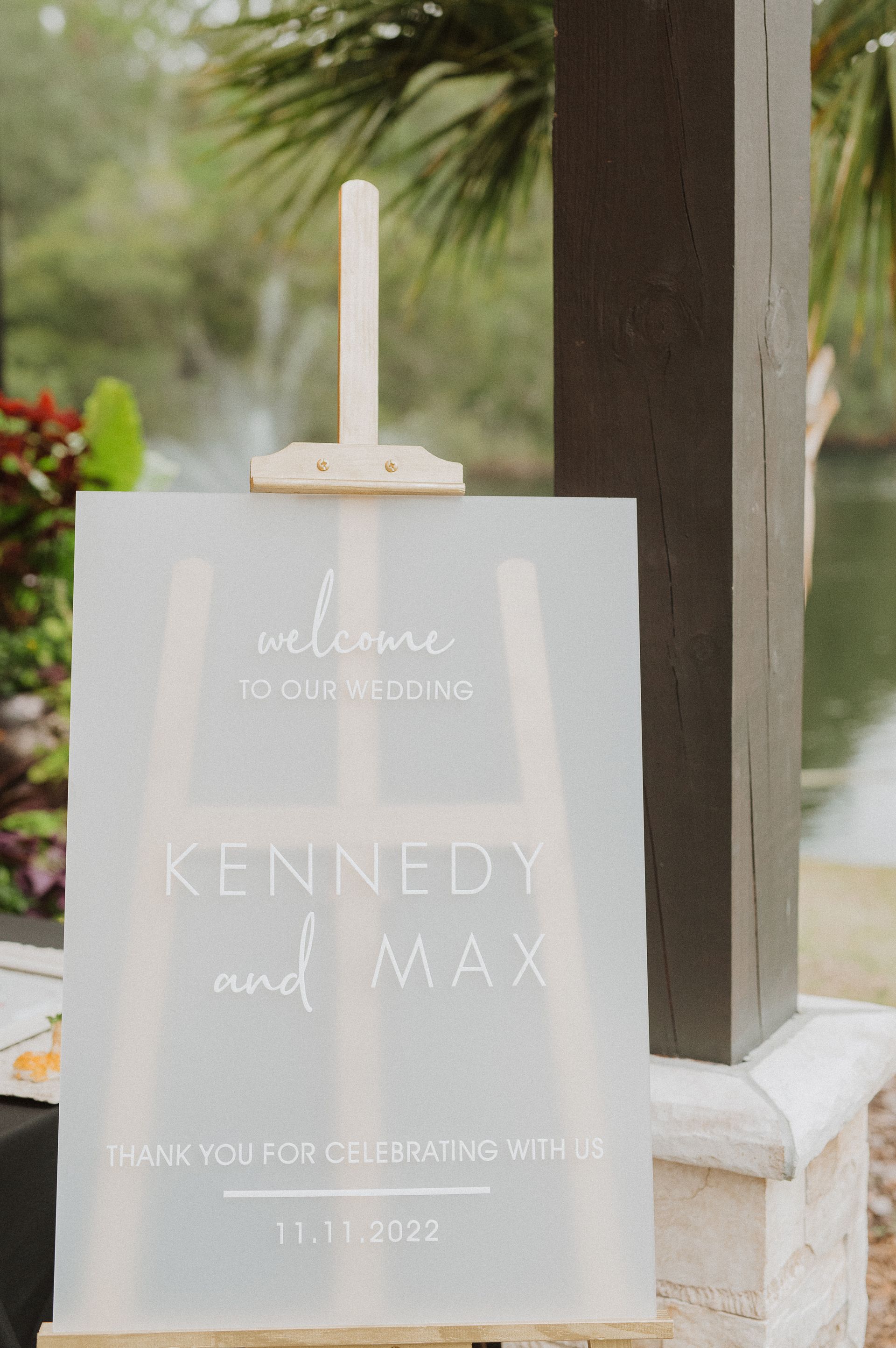 White memorial sign with “Kennedy and Max” on a stand outdoors near greenery and columns