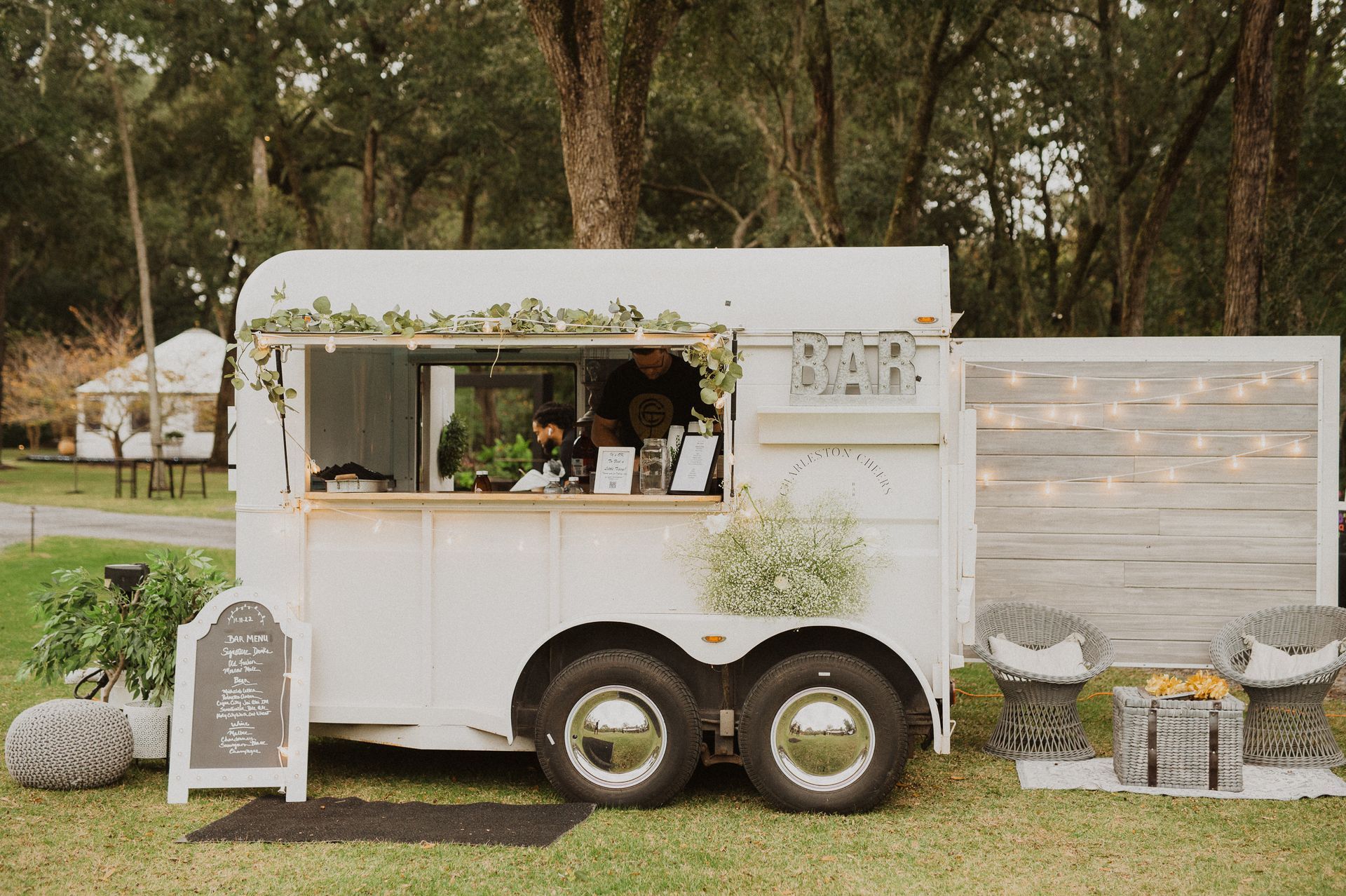 White food truck at an outdoor event, with chalkboard sign and floral decor, parked on grass among trees