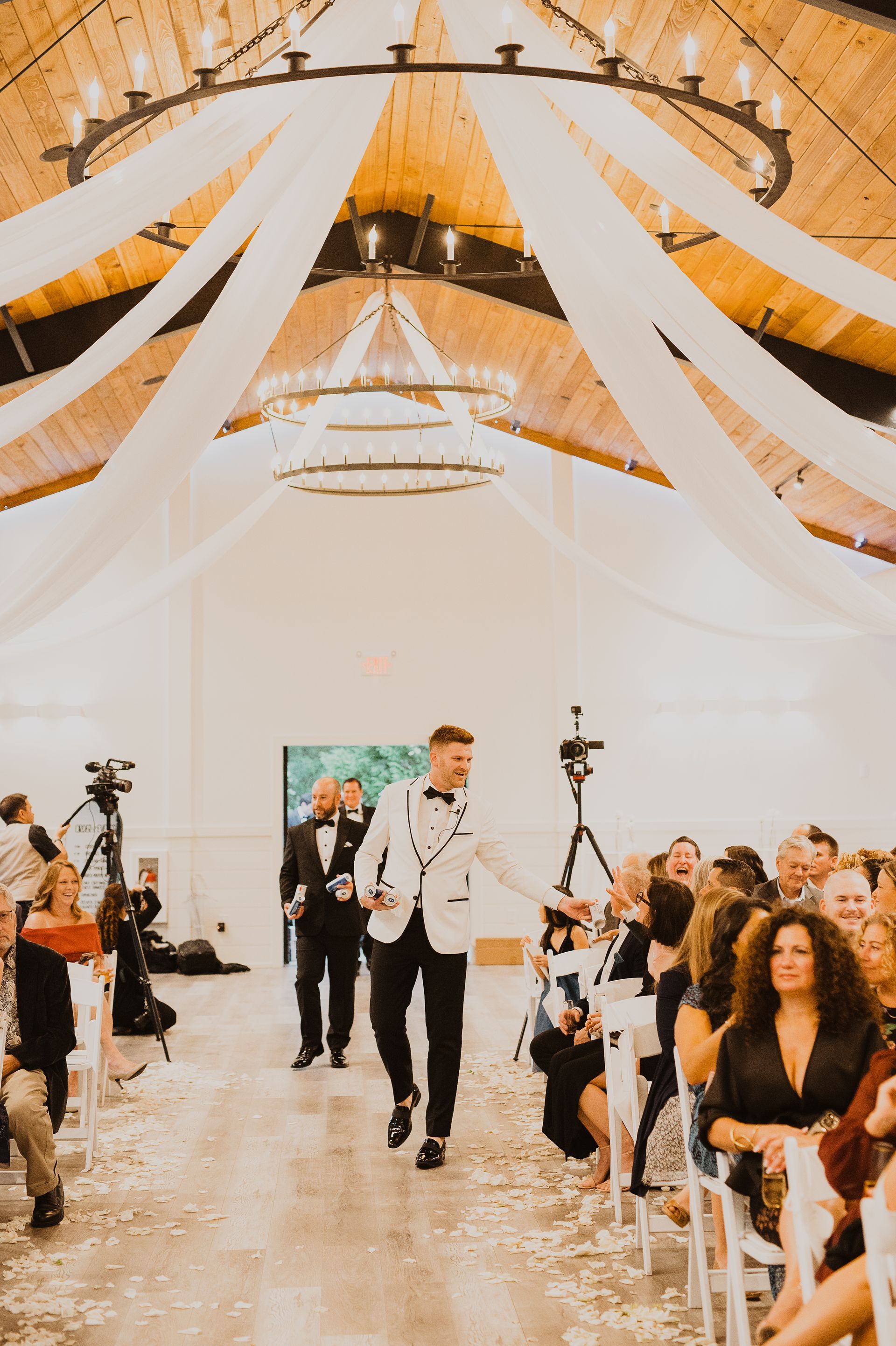 Wedding aisle with guests seated under white drapes and warm wood ceiling, two men in white suits walking вперед.