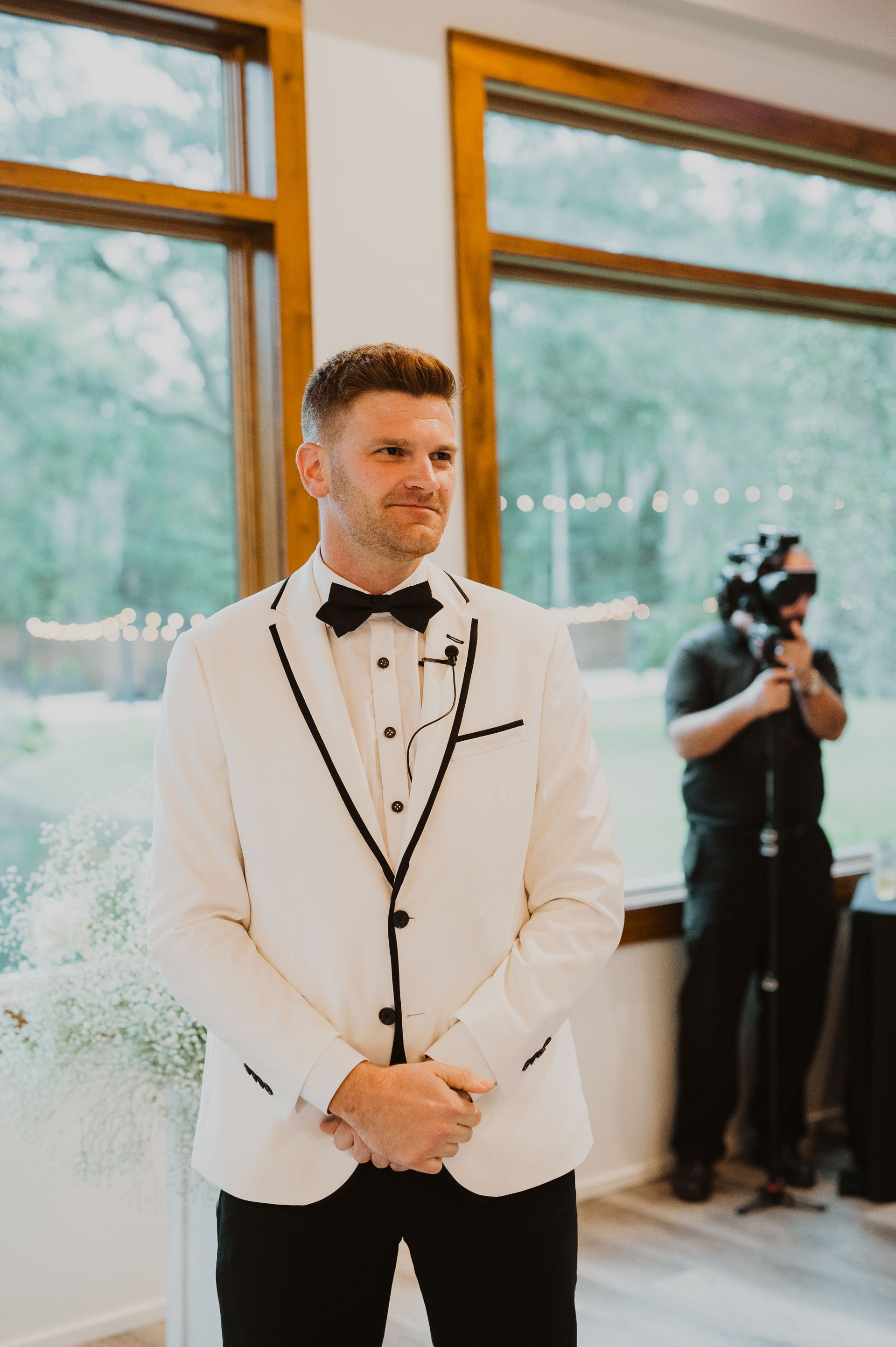Man in a white tuxedo jacket stands by large windows at a wedding venue while a photographer works nearby