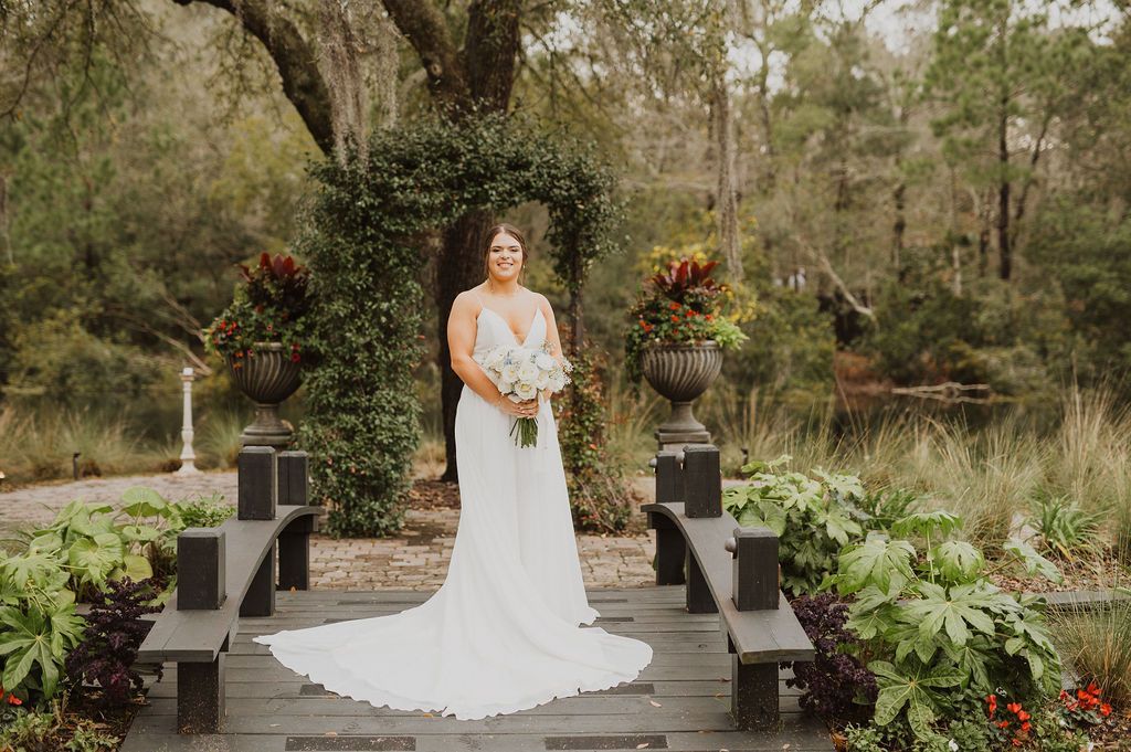 Bride in a white gown holding a bouquet on an outdoor garden walkway, framed by floral arrangements