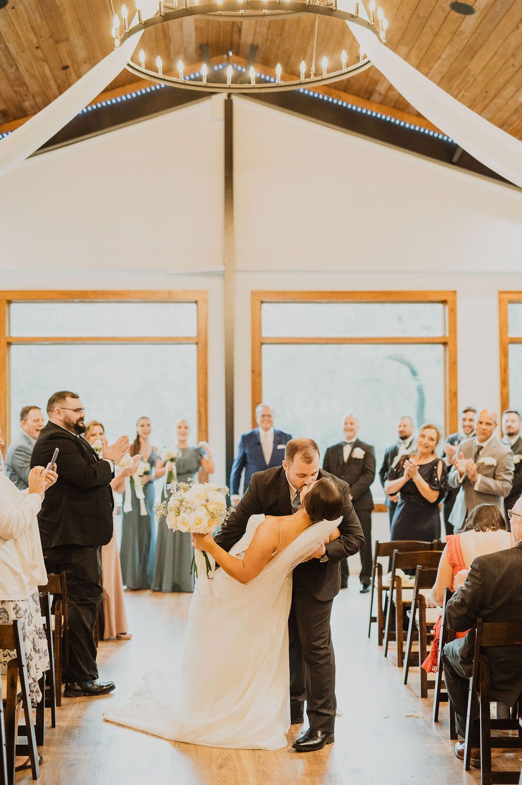 Bride and groom kiss at wedding ceremony in a bright church, guests applauding nearby