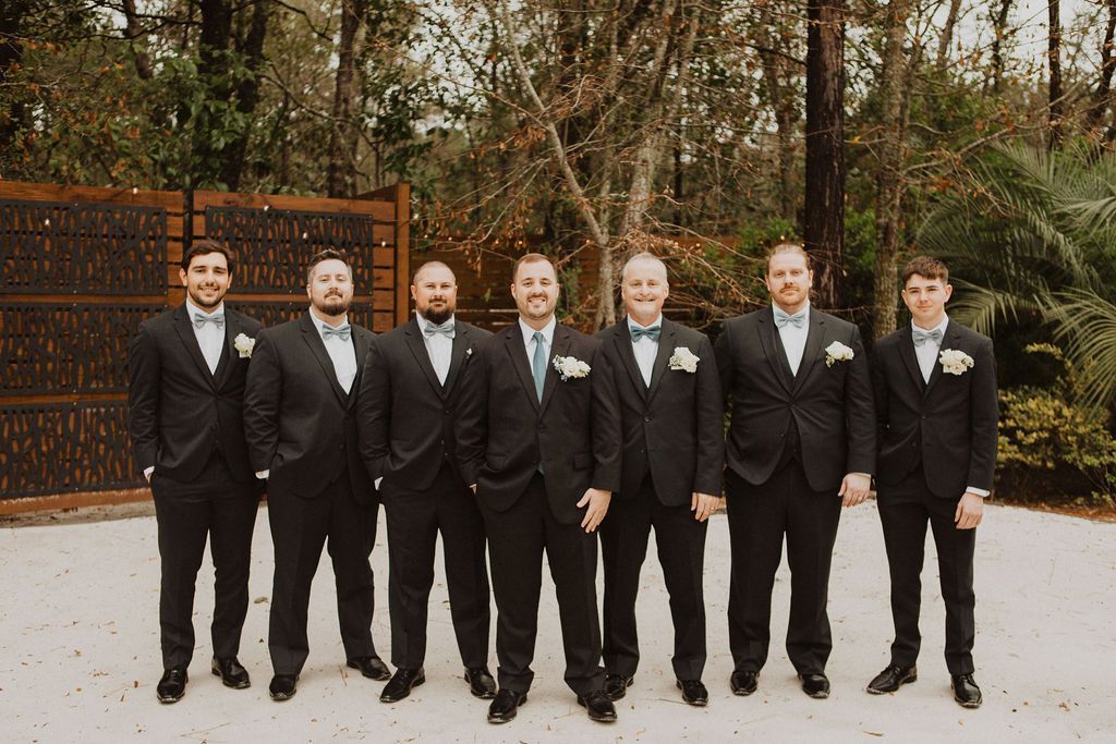 Seven men in black suits stand outdoors in a formal group portrait.