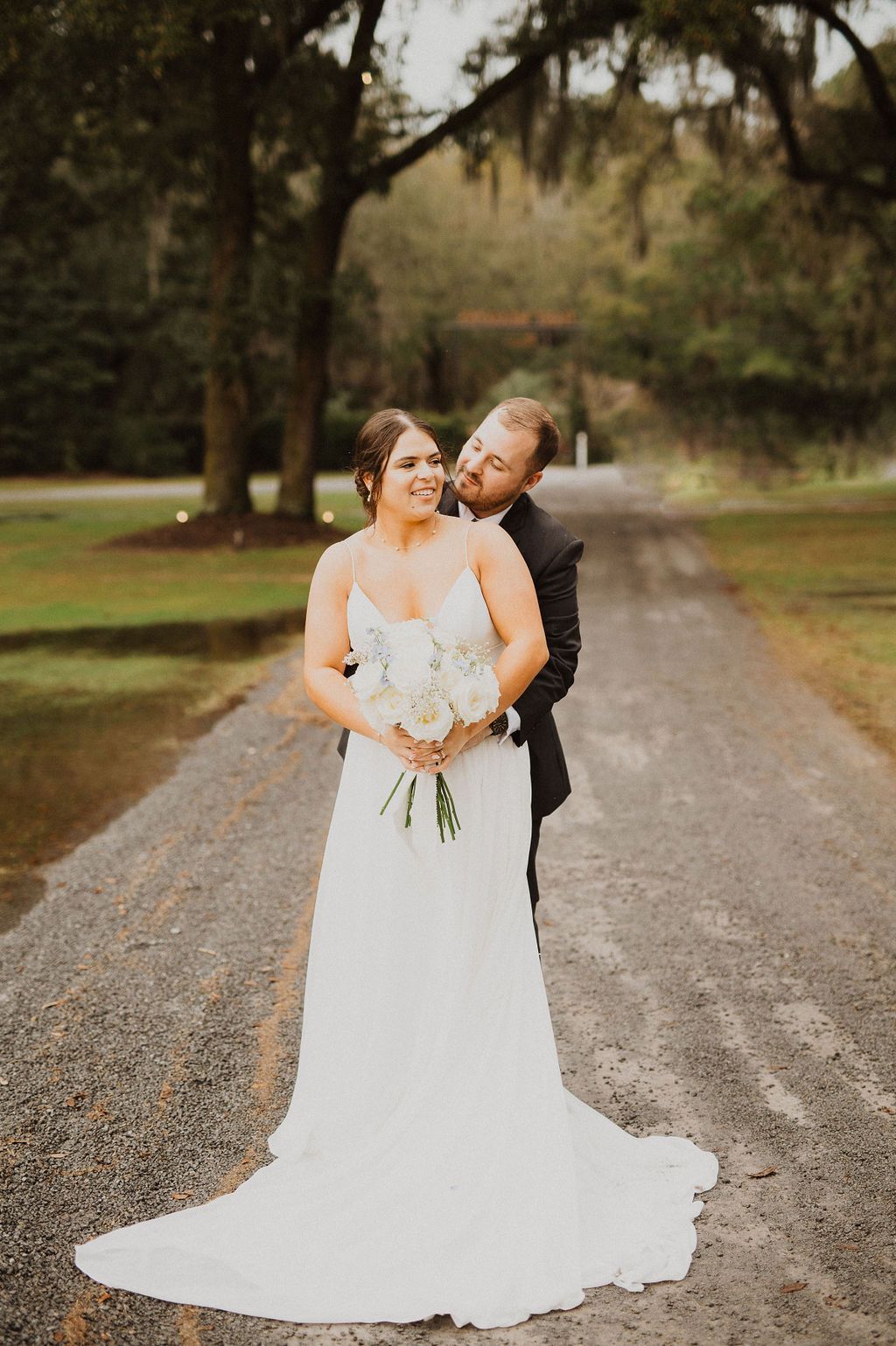 Bride and groom embracing on a tree-lined gravel road, bride in white gown holding bouquet