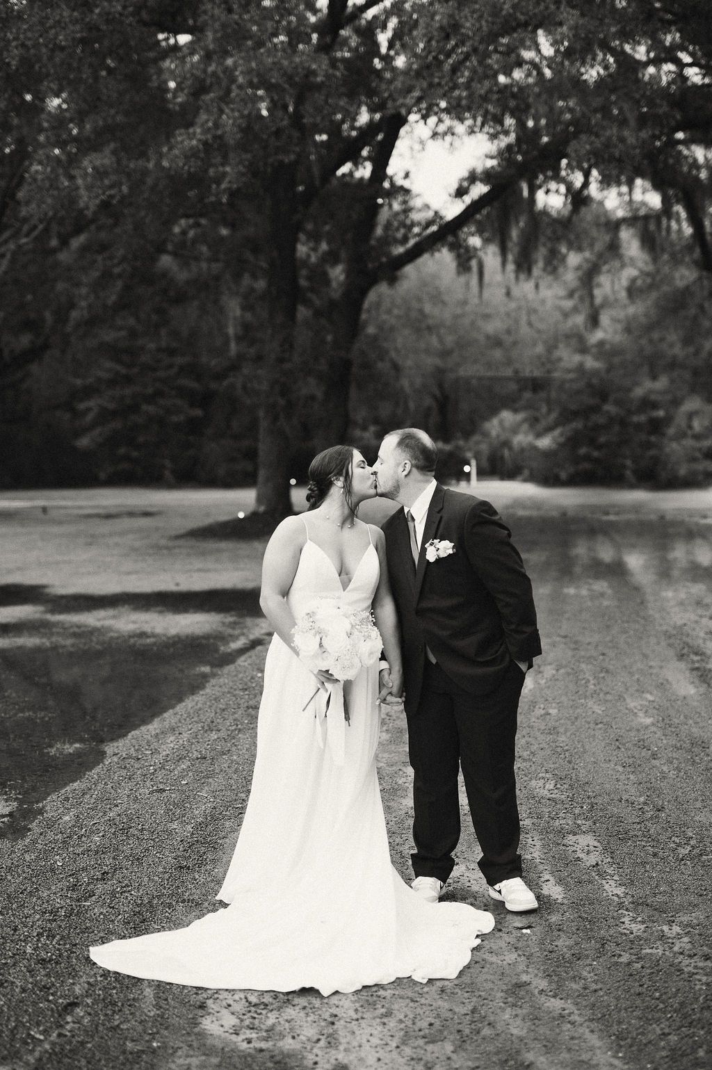 Bride and groom kissing on a gravel path under trees, in a black-and-white wedding photo