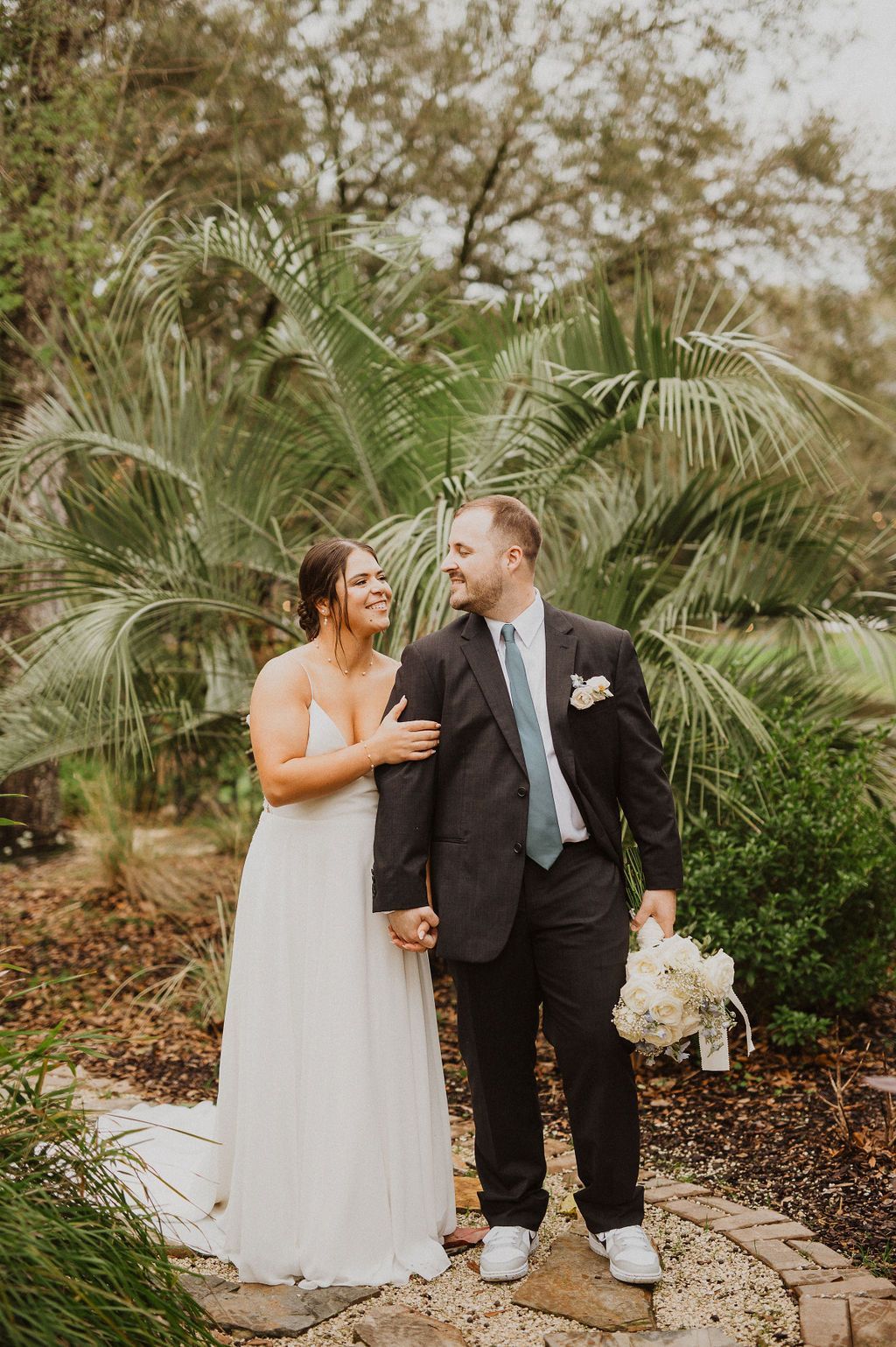 Bride and groom standing together in a garden with palm trees, smiling and holding hands.