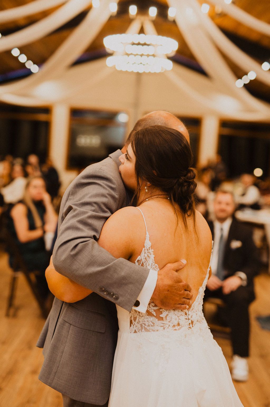 Couple sharing a hug and kiss at a wedding reception, with guests and chandeliers in the background