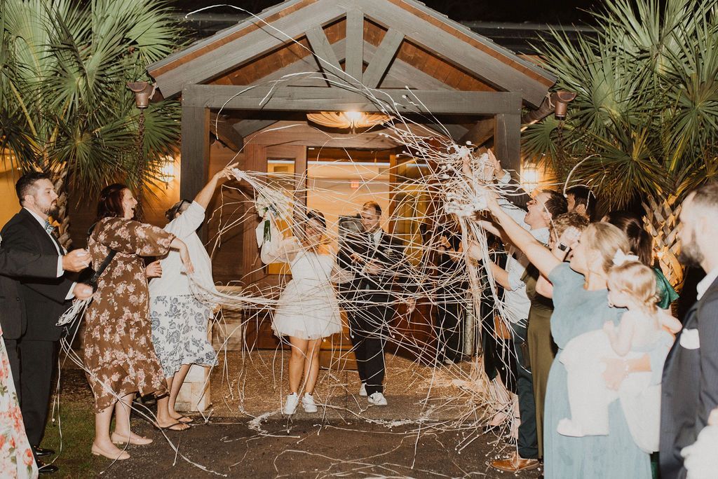 Wedding guests toss confetti as the bride and groom exit a decorated venue at night.