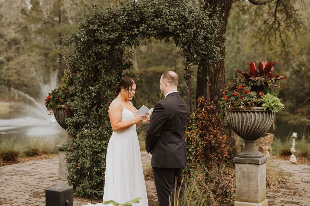 Couple exchanging rings under a floral arch in a garden by the water