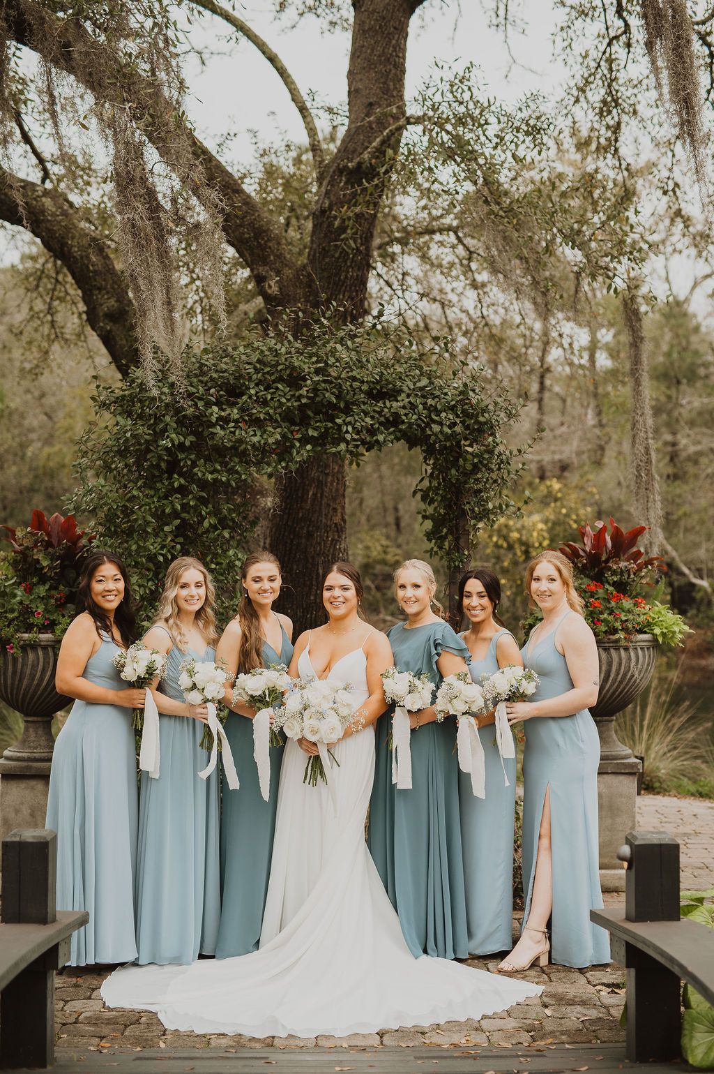 Wedding party posing outdoors in light blue dresses beside the bride in a white gown under trees.