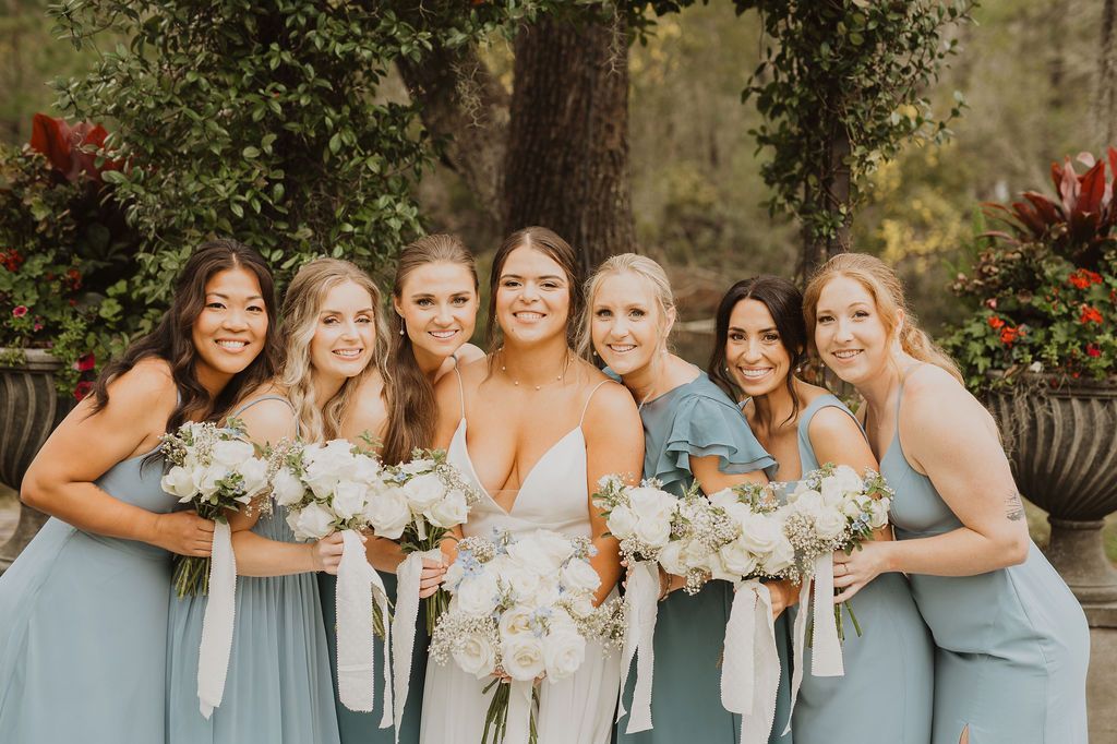 Seven bridesmaids in sage dresses holding white bouquets in a garden, smiling together.