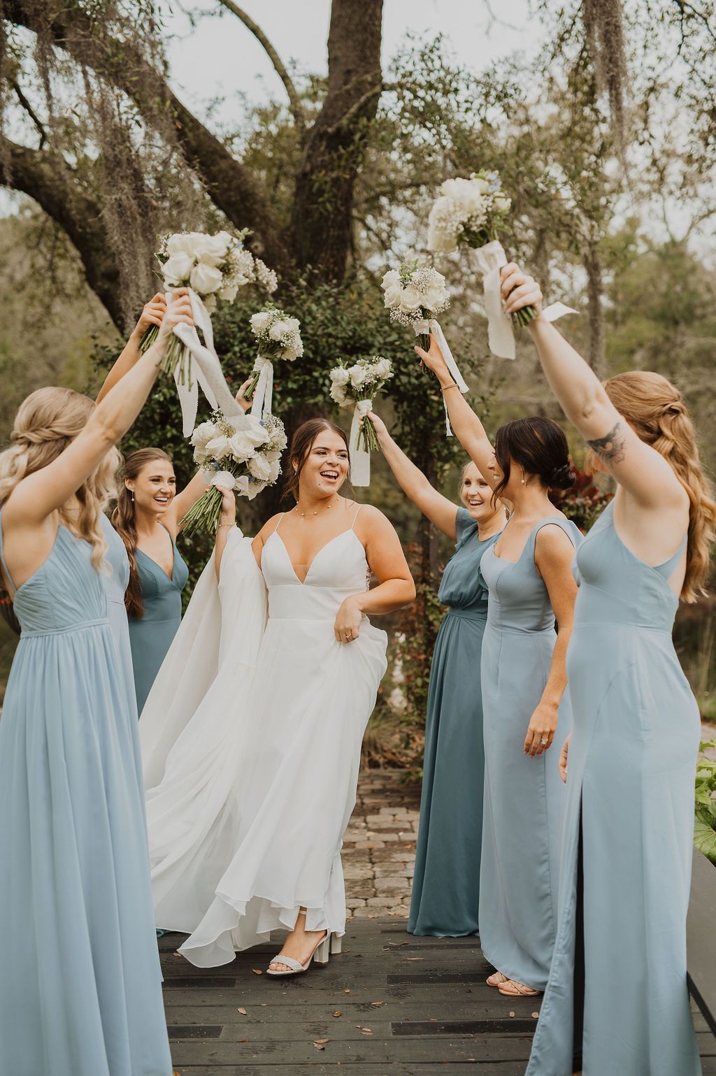 Bridal party in blue dresses raising bouquets around a bride in white on a wooden bridge outdoors
