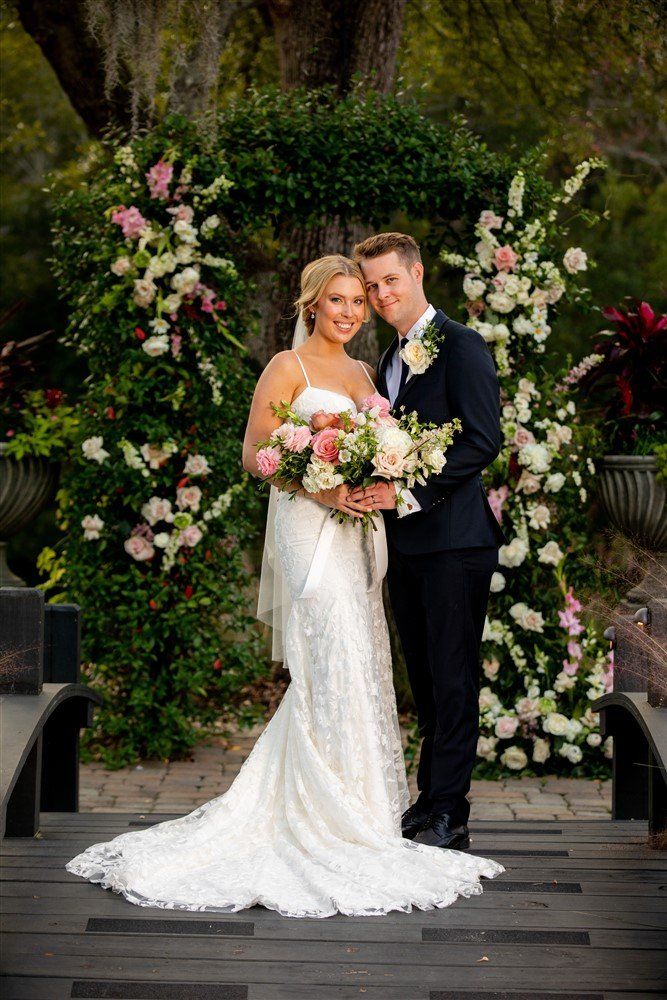 Wedding couple standing together outdoors, bride in white gown holding bouquet, groom in dark suit amid floral arch