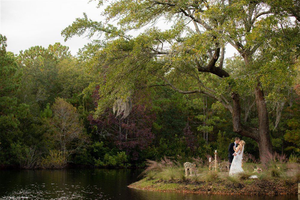 Bride in white standing by a riverbank under large trees, with lush green foliage in the background