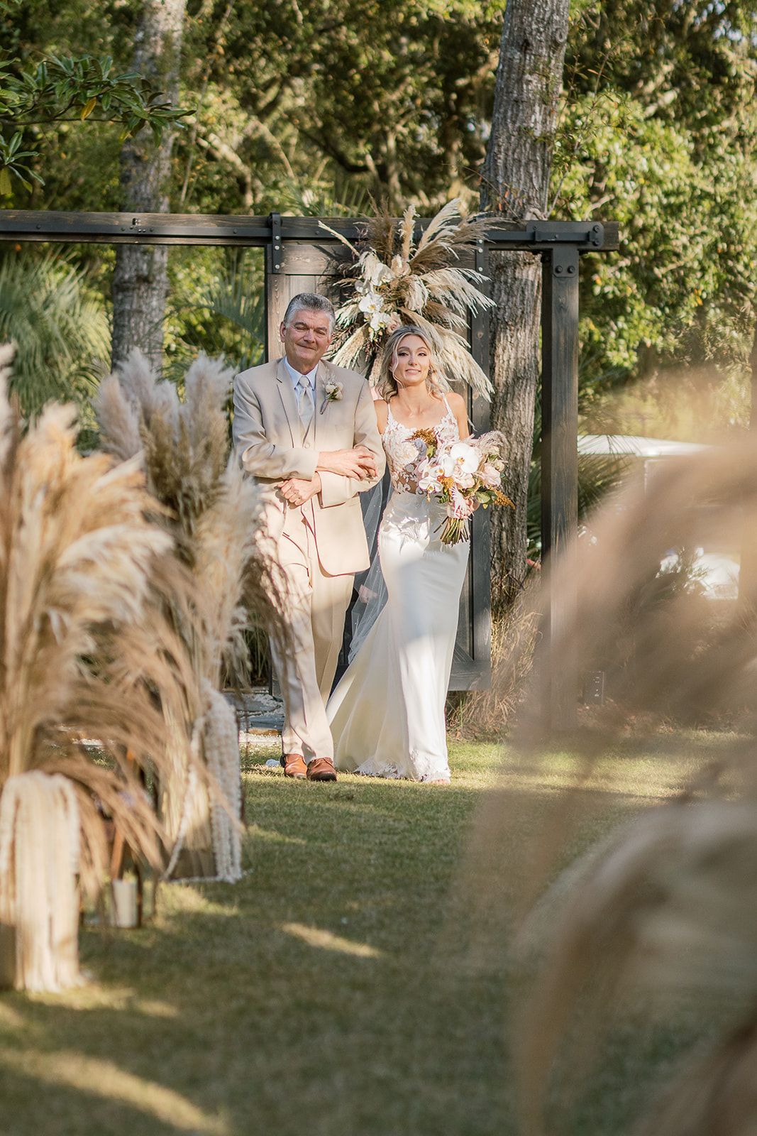 Couple standing together in a garden ceremony aisle under a floral arch with pampas grass in the foreground