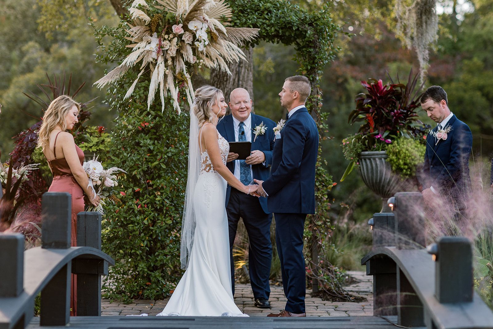 Wedding couple exchanging vows outdoors beside floral arrangements and an officiant