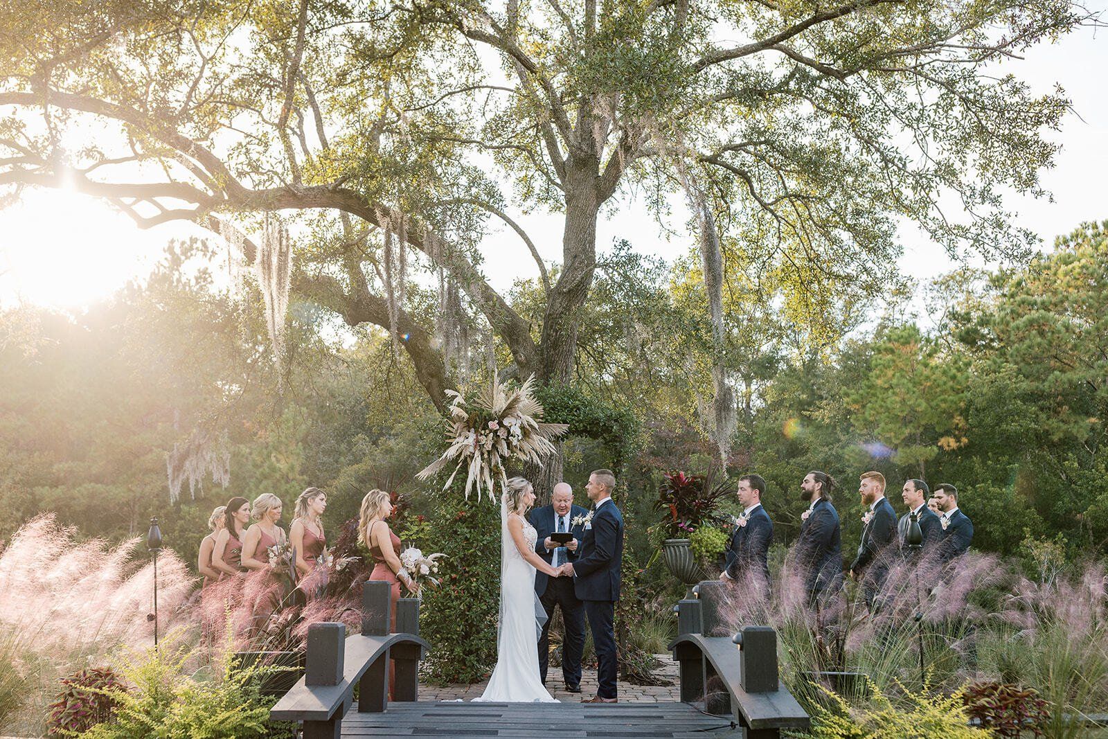 Outdoor wedding ceremony under trees at sunset, with couple and wedding party on a small stage