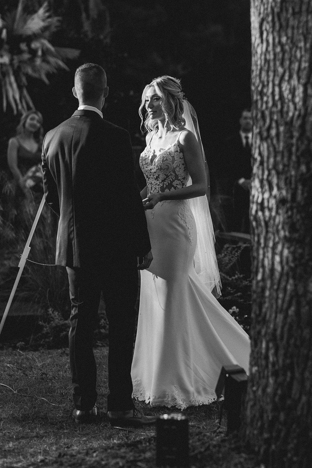 A couple in formal attire standing outdoors beside a tree at night.