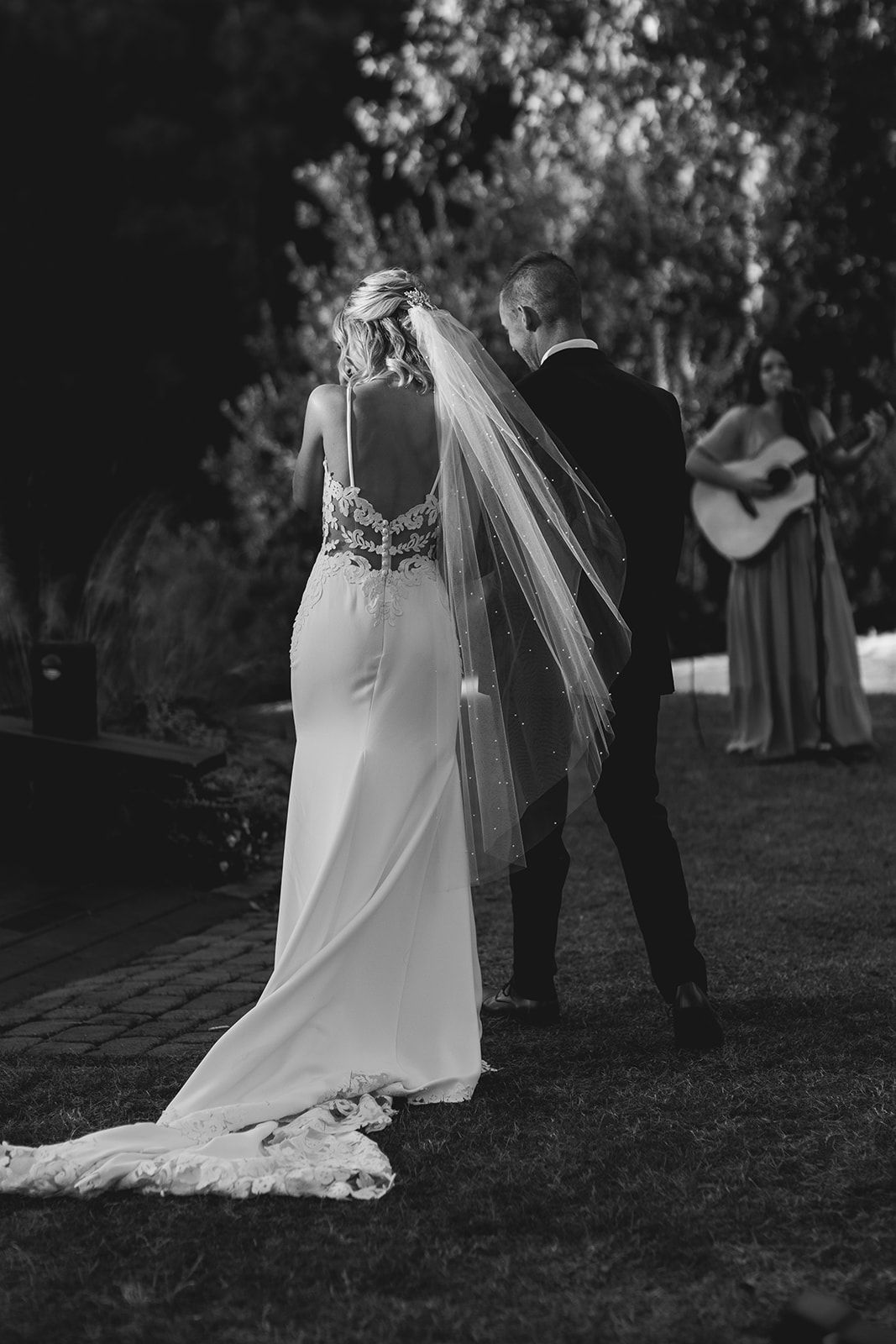 Bride and groom embrace in a garden during an outdoor wedding, with a guitarist in the background.
