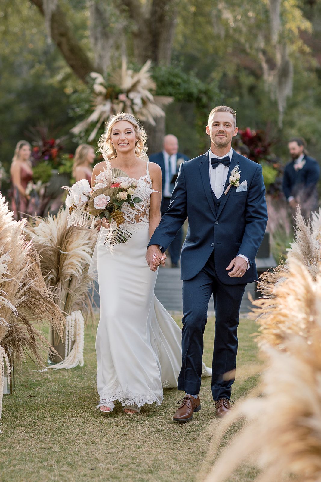 Bride and groom walking down an outdoor aisle with floral decorations, holding a bouquet and wearing formal attire