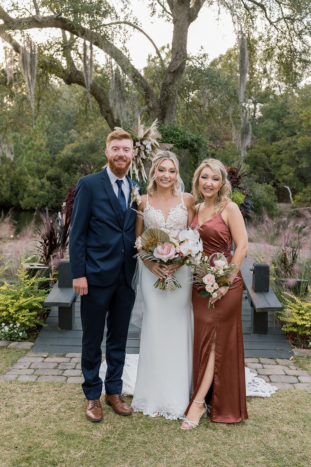 Three people posing outdoors at a wedding, with the bride in white and two attendants in dark and rust dresses.