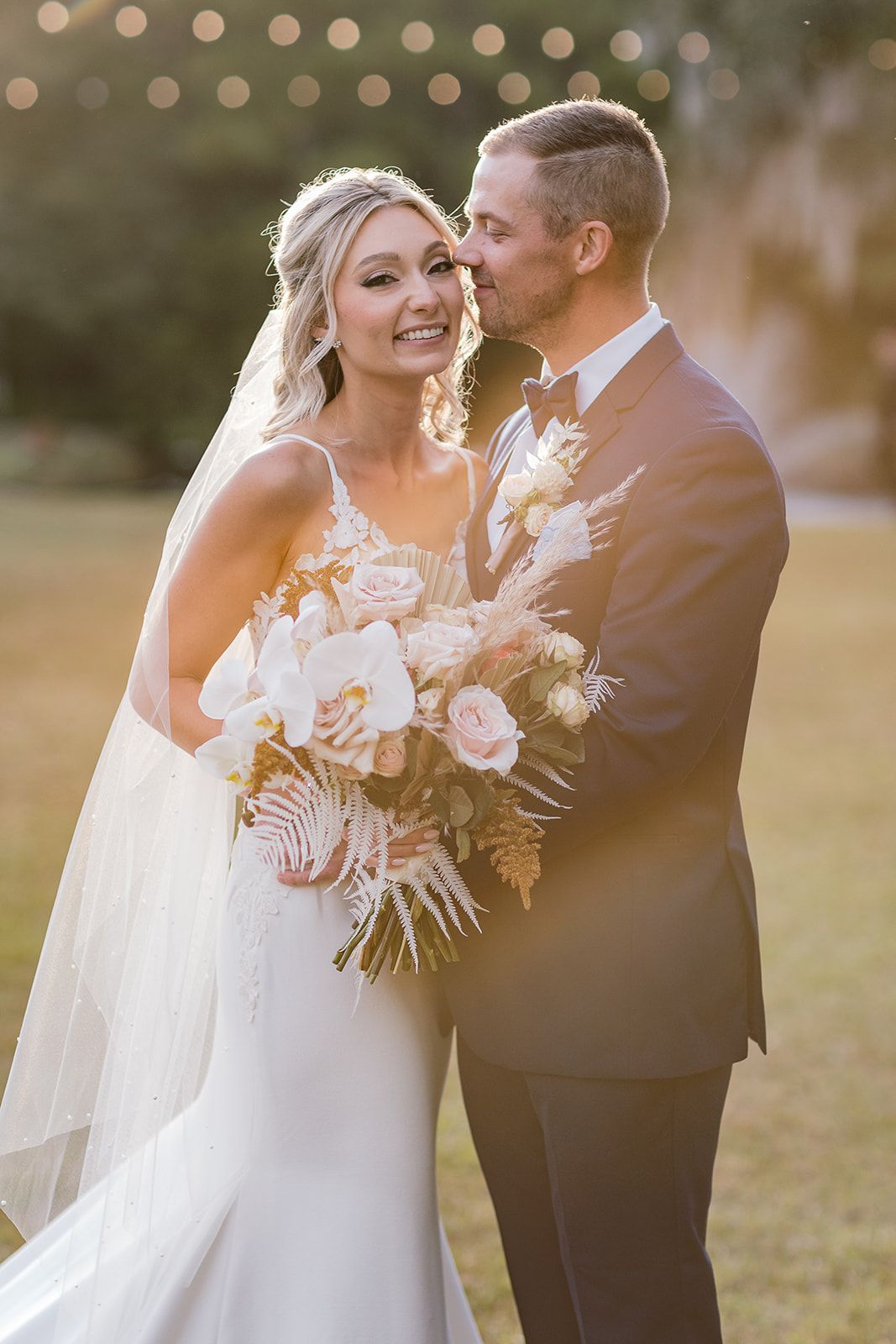 Bride and groom standing together outdoors at sunset, smiling and holding a white floral bouquet