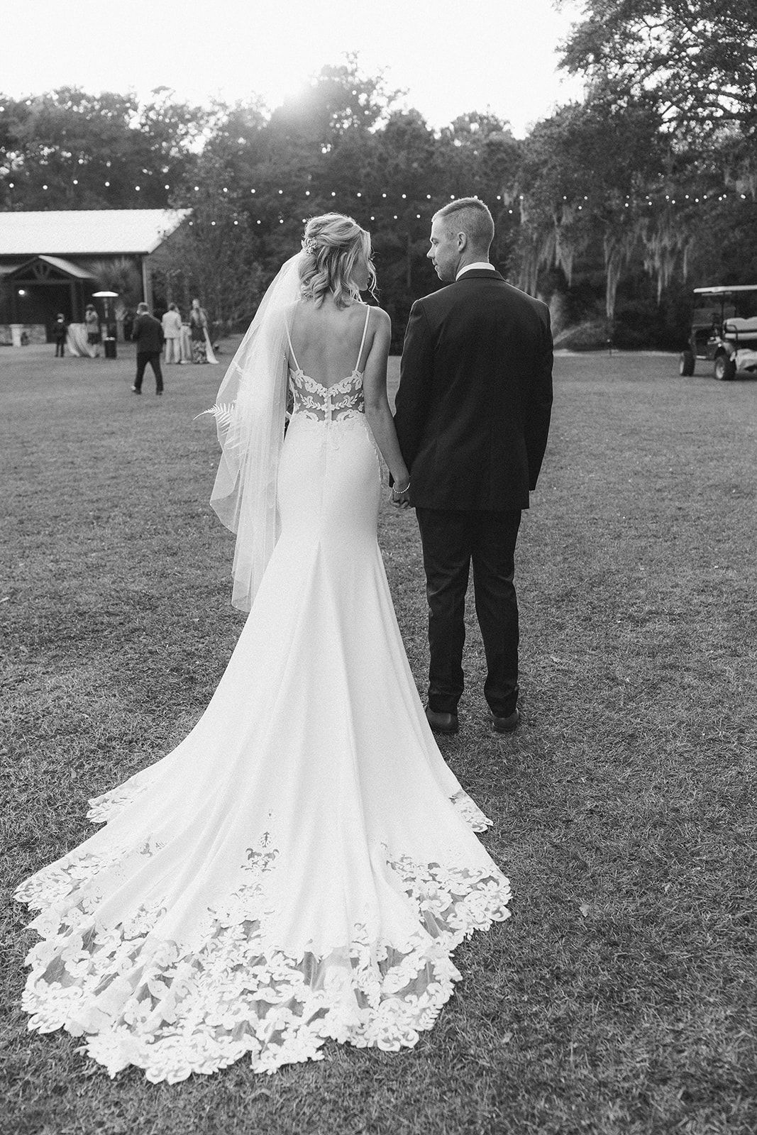 Bride and groom standing together outdoors in a grassy field, bride in a long lace wedding gown and veil.