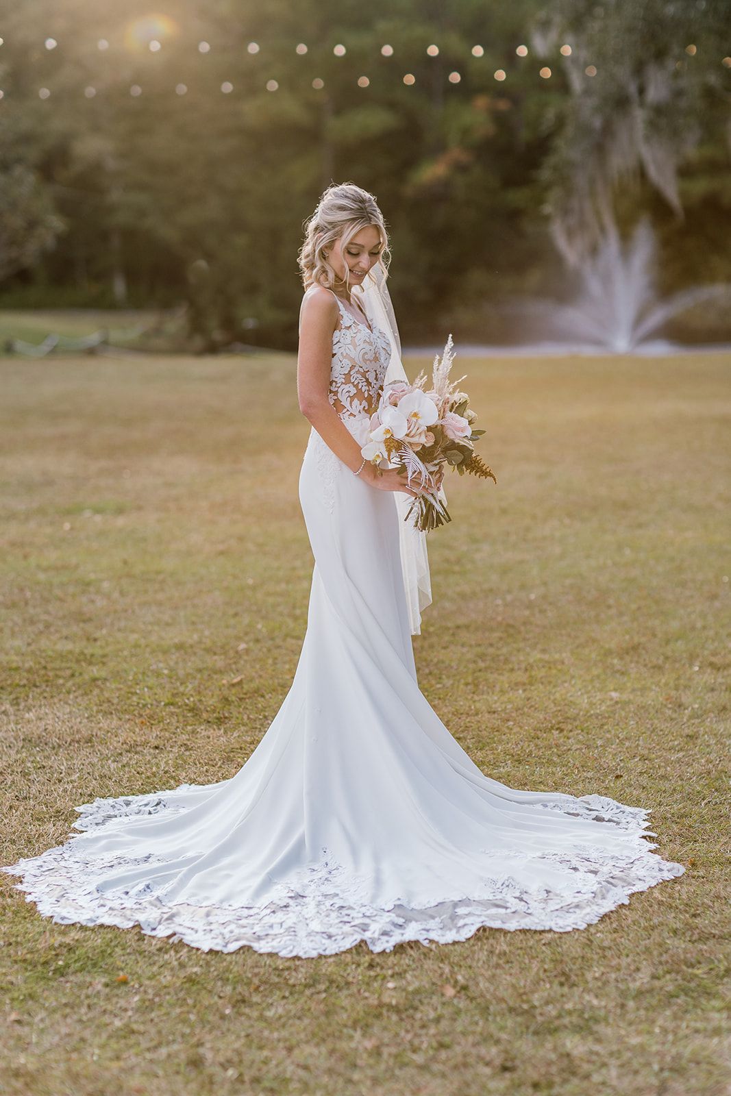 Bride in a white wedding gown holding a bouquet on a grassy lawn at sunset