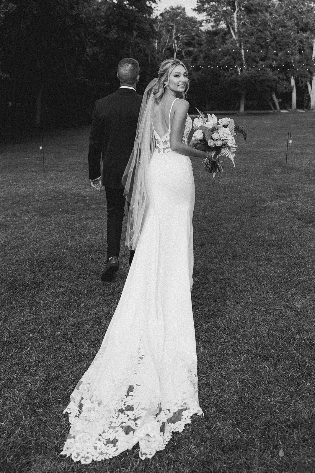 Bride in a long white gown holding a bouquet, standing outdoors in a grassy area with a man behind her.