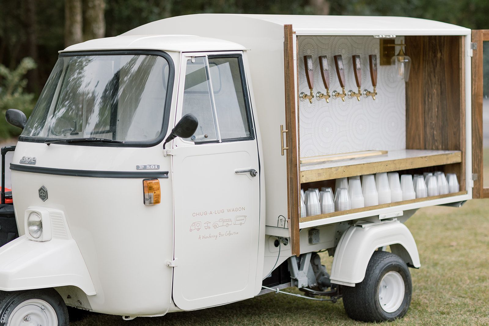 White coffee cart trike with serving window, drink taps, and shelves of cups on a grassy field
