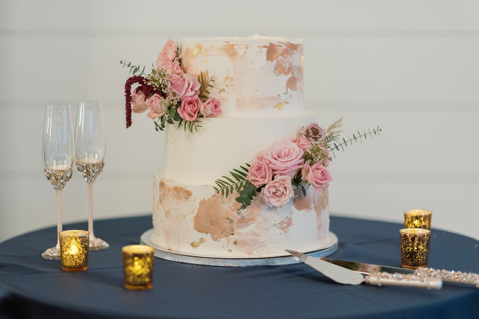 Two-tier white wedding cake with pink flowers, champagne flutes, and gold candle holders on a navy tablecloth